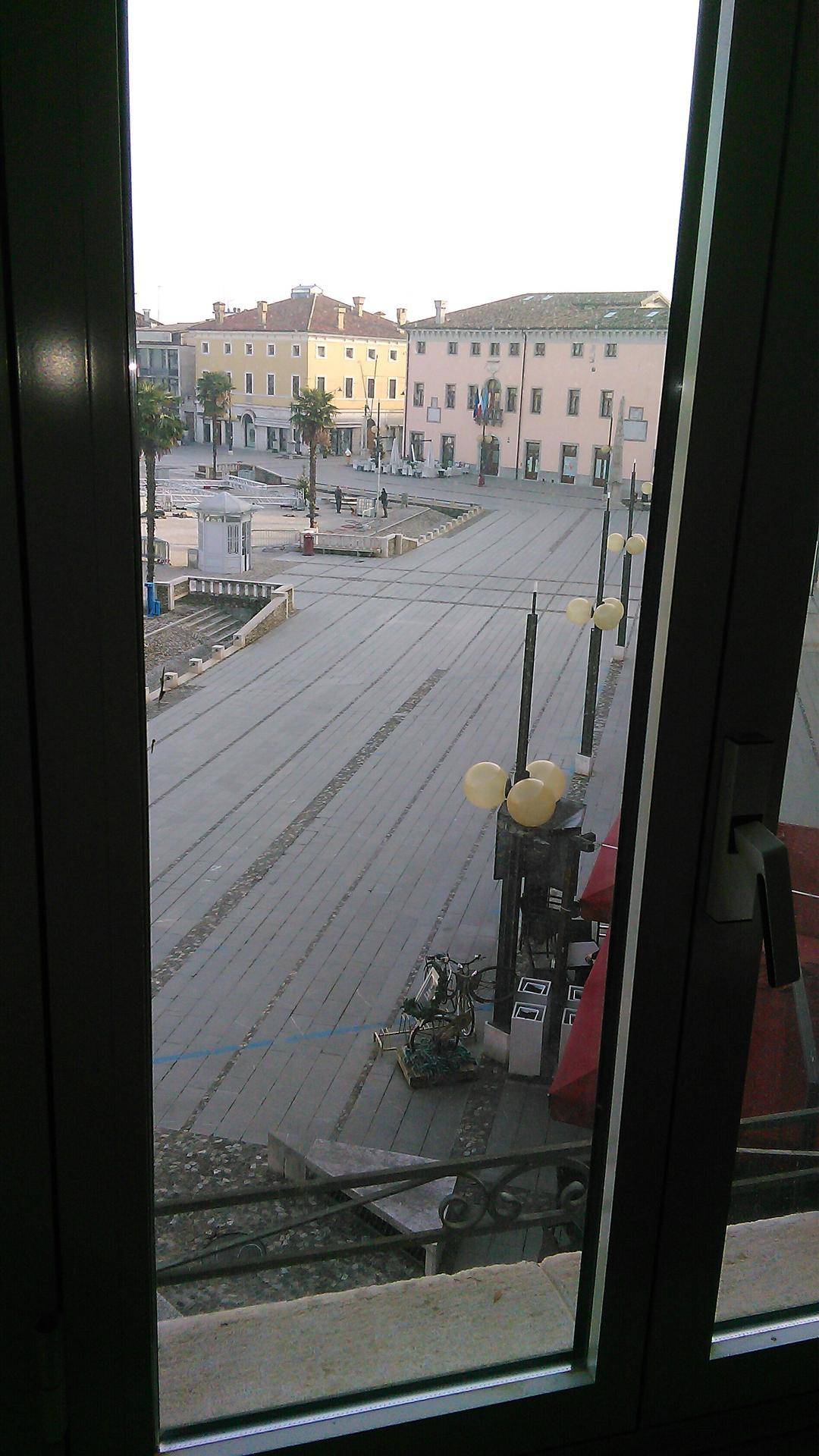 Blick aus einem Fenster auf einen belebten Stadtplatz mit Gebäuden und Straßenlaternen.
