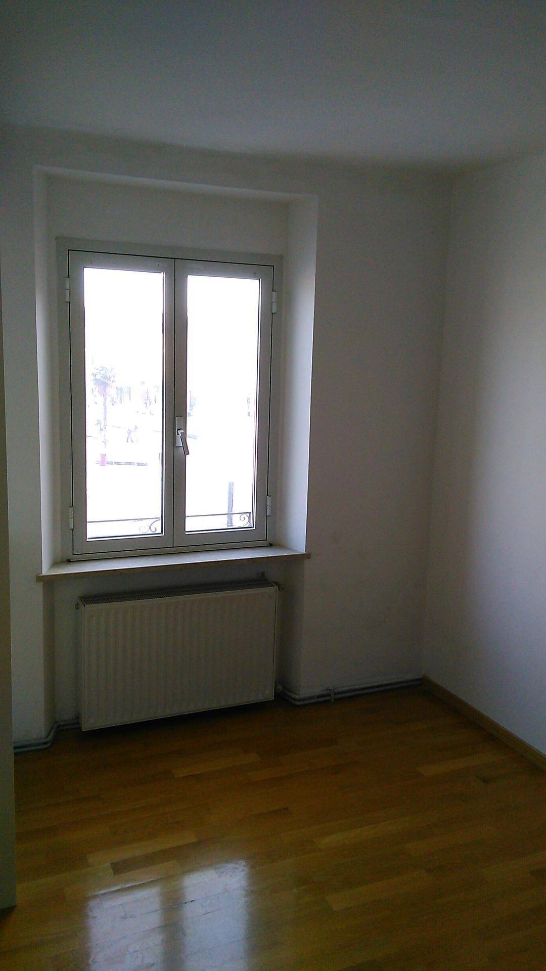 Heller, leerer Raum mit Holzboden und einem großen Fenster, das viel Tageslicht hereinlässt.