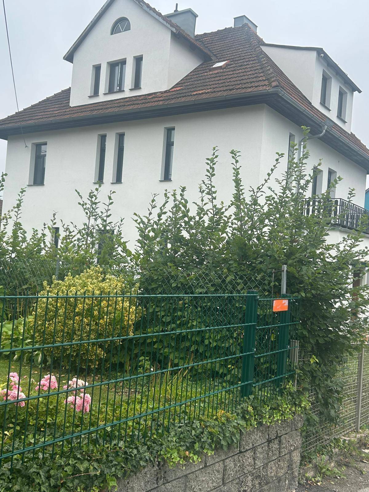 Wohnhausfassade in Weiß mit Satteldach, umgeben von einem grünen Zaun und blühenden Pflanzen.