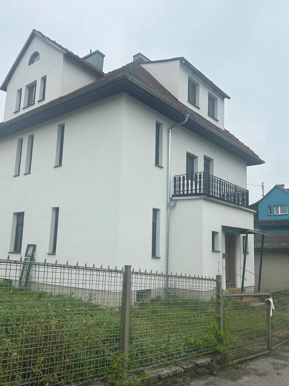 Seitenansicht eines weißen Wohnhauses mit kleinem Balkon und dunklem Geländer.