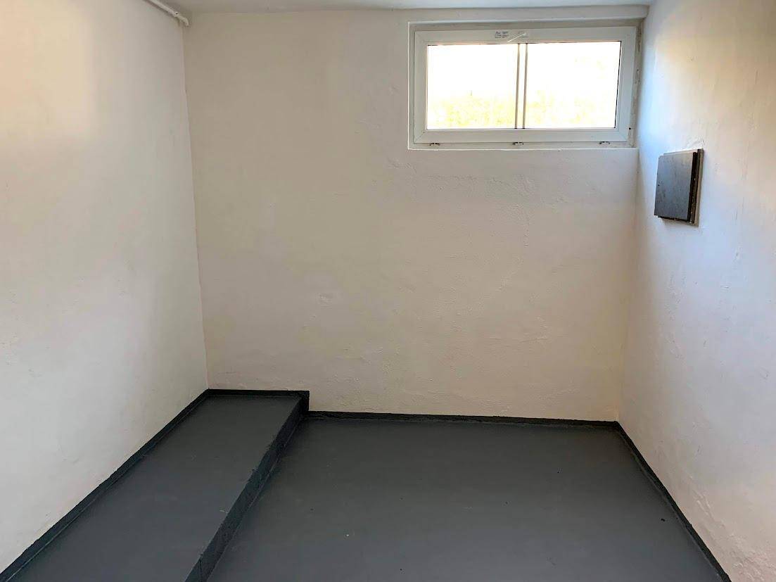 Kleiner Lagerraum mit grauem Boden, weißen Wänden und einem hochgelegenen Fenster, mit erhöhter Ablagefläche.