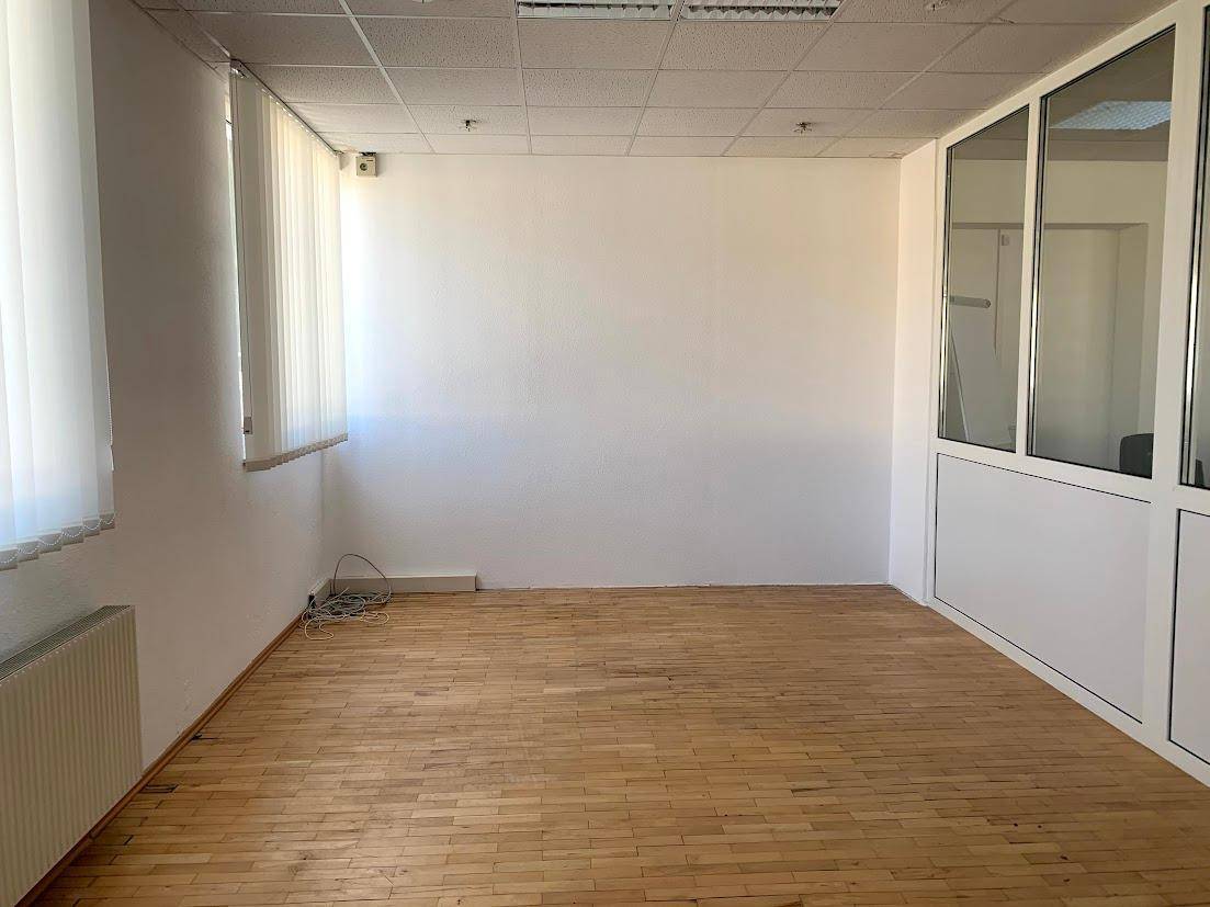Leeres Büro mit hellem Holzparkett, weißen Wänden und Fenster mit Jalousien, abgetrennt durch eine Glaswand.