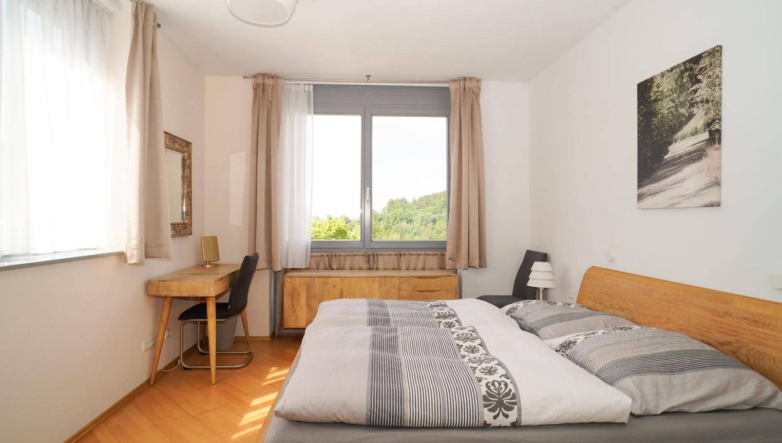 Helles Schlafzimmer mit Doppelbett, Holzmöbeln und großem Fenster mit Grünblick.
