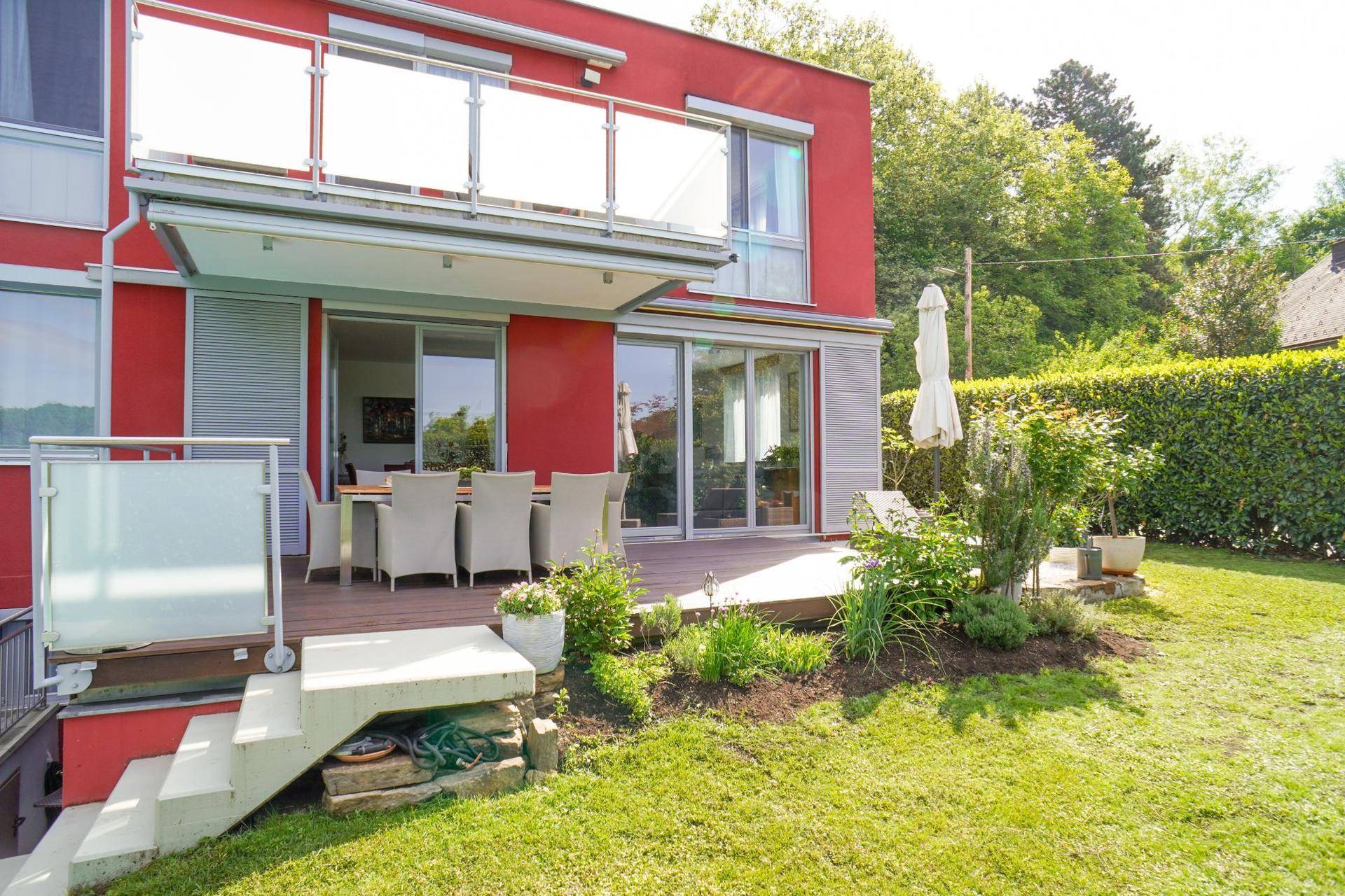 Rote Hausfassade mit großer Holzterrasse, Garten und Zugang zum Untergeschoss.