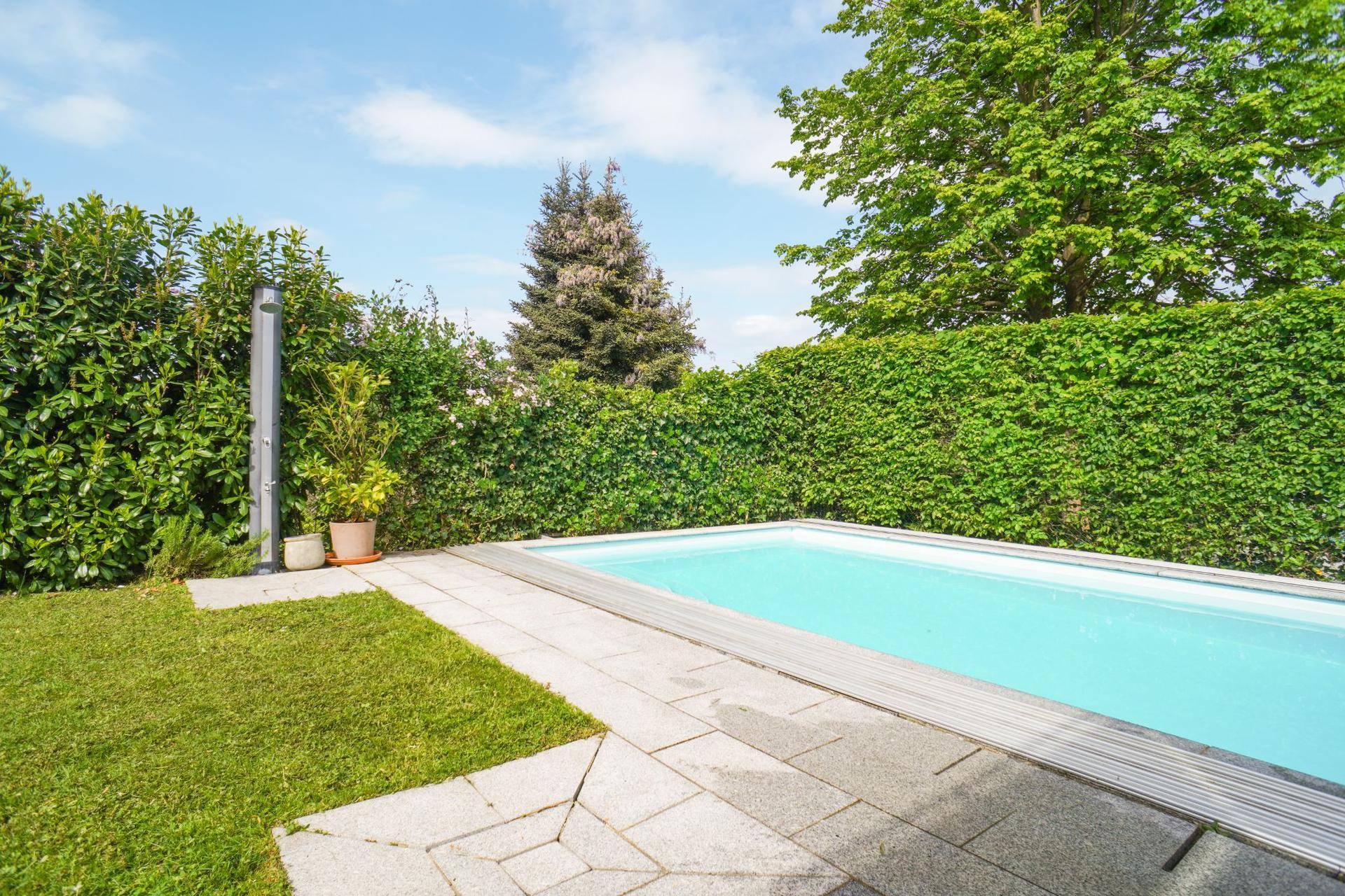 Außenpool mit gepflastertem Bereich, umgeben von Rasen und einer grünen Hecke.