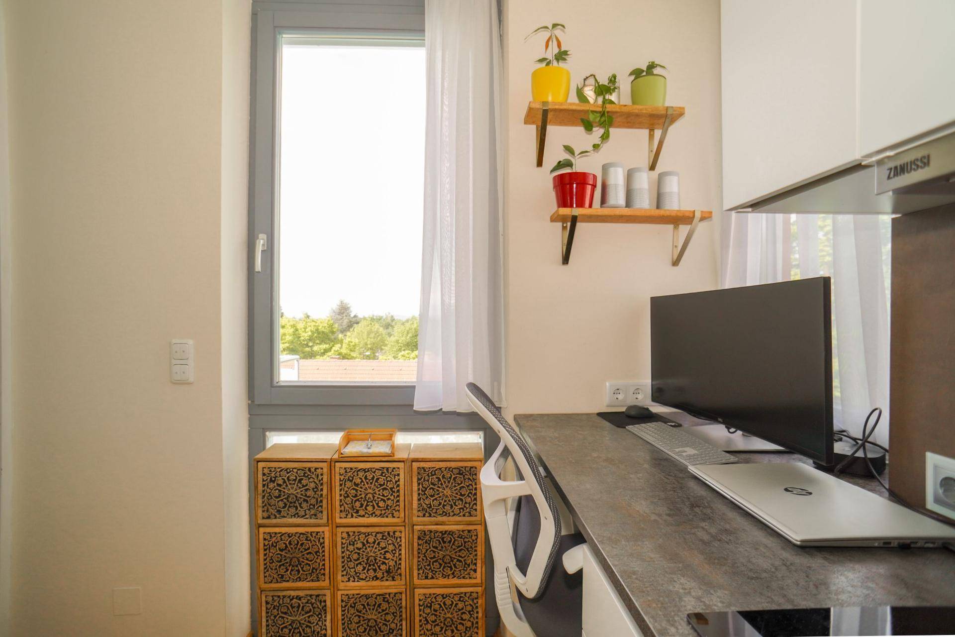 Heller Arbeitsplatz mit Schreibtisch, Monitor und Fenster mit Blick ins Grüne.