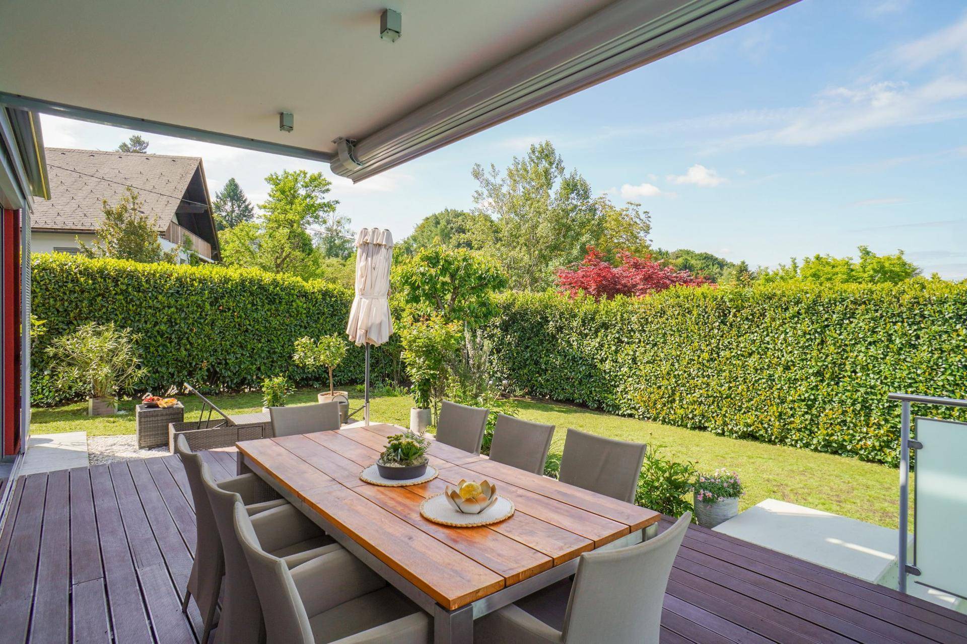 Überdachte Holzterrasse mit großem Esstisch und Stühlen, umgeben von viel Grün.