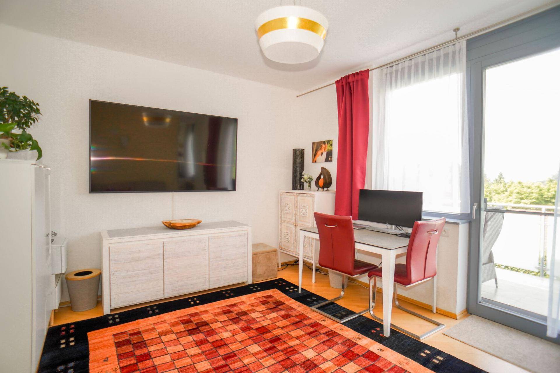 Helles Arbeitszimmer mit großem Fernseher, Schreibtisch und Zugang zum Balkon.