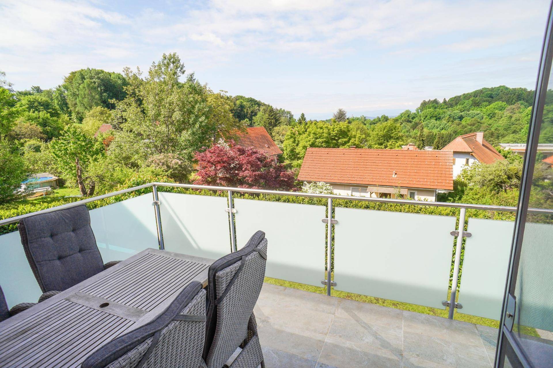Möblierter Balkon mit Glasgeländer und weitem Blick über die grüne Umgebung.