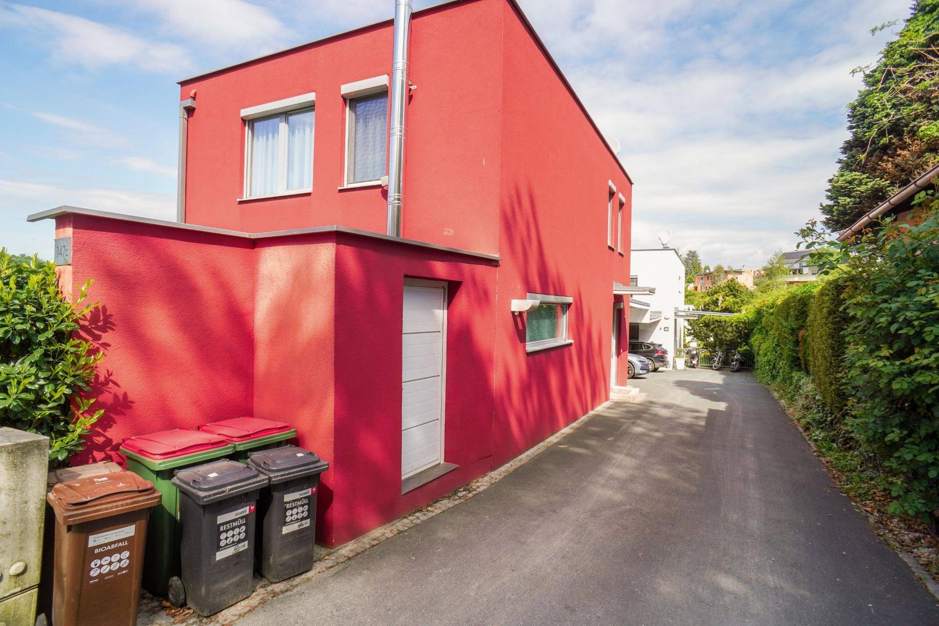 Seitenansicht des roten Hauses mit Mülltonnen und Zufahrtsweg.
