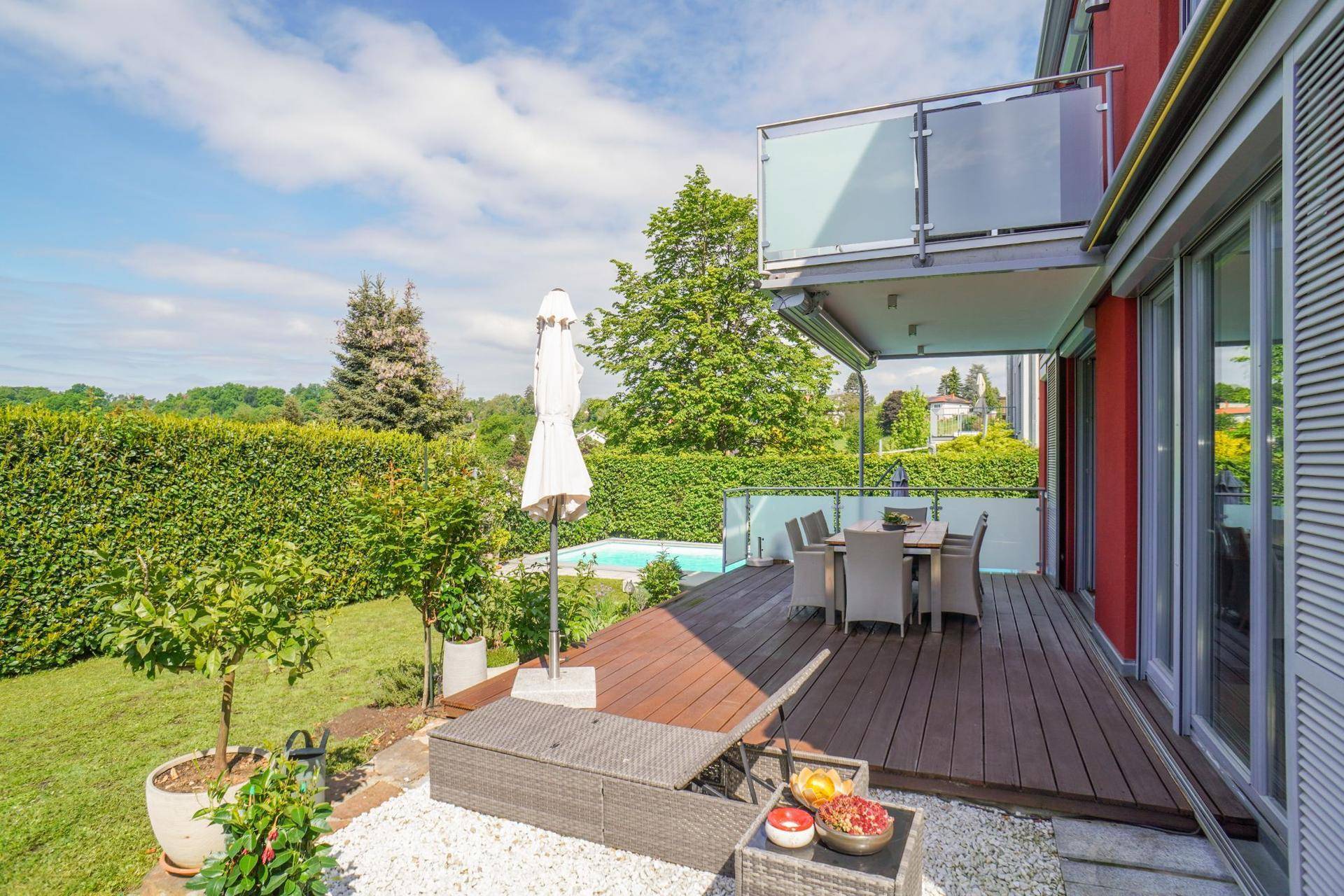 Geräumige Holzterrasse mit Sonnenschirm, Liegestuhl und Blick auf den Garten und Pool.