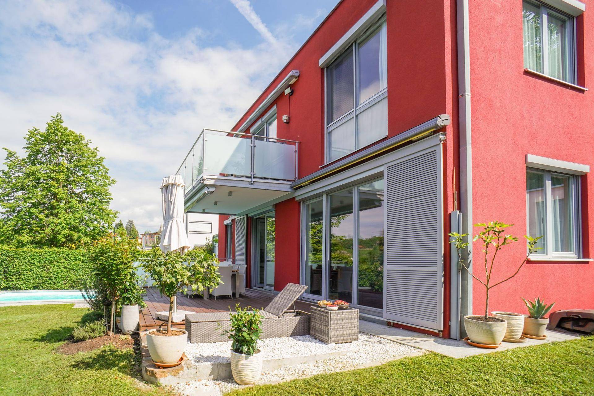 Rote Hausfassade mit großer Holzterrasse, Liegestuhl und gepflegtem Garten.