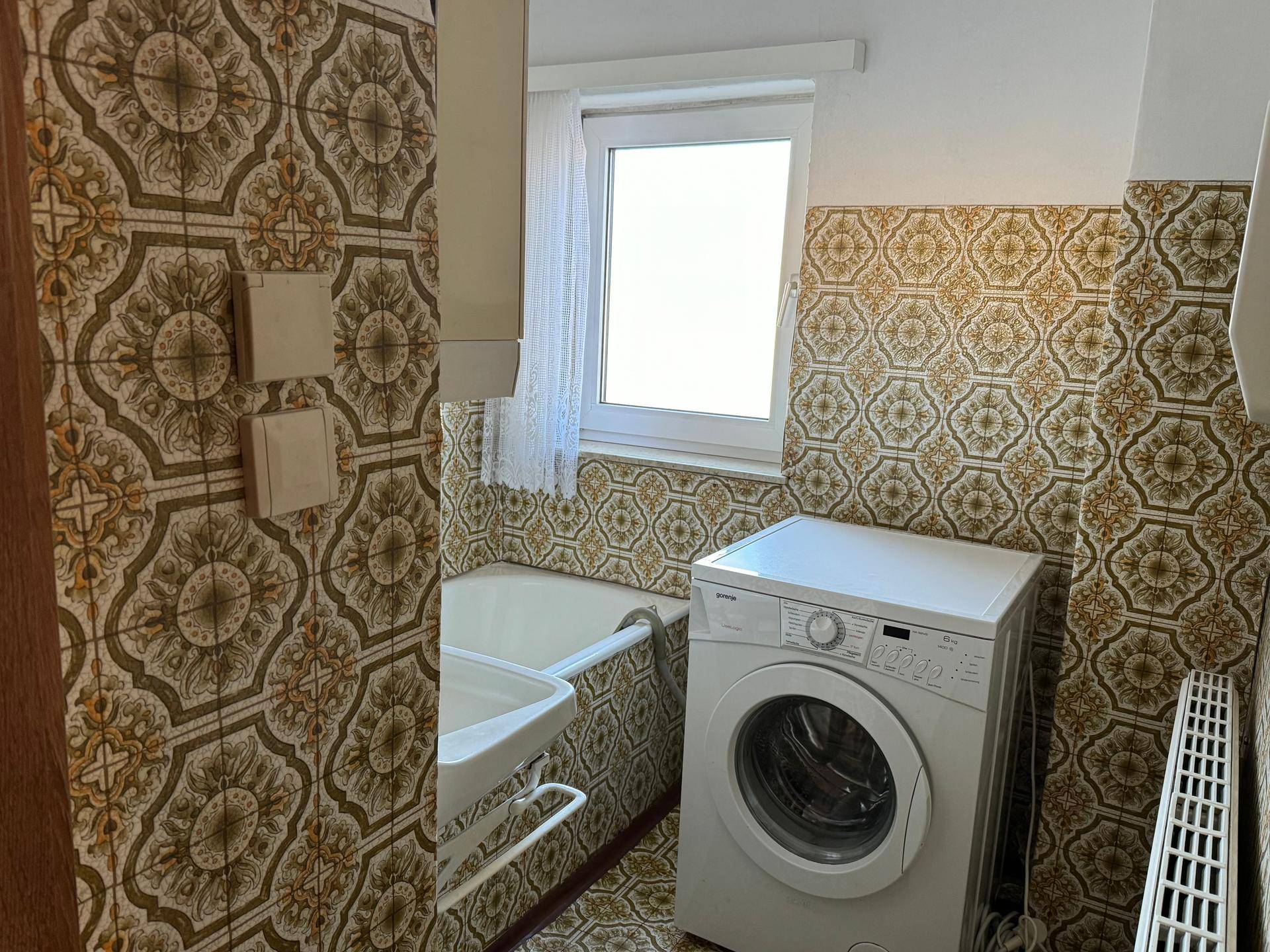 Badezimmer im Stil der 80er Jahre mit gemusterten Fliesen, Badewanne und Waschmaschine.