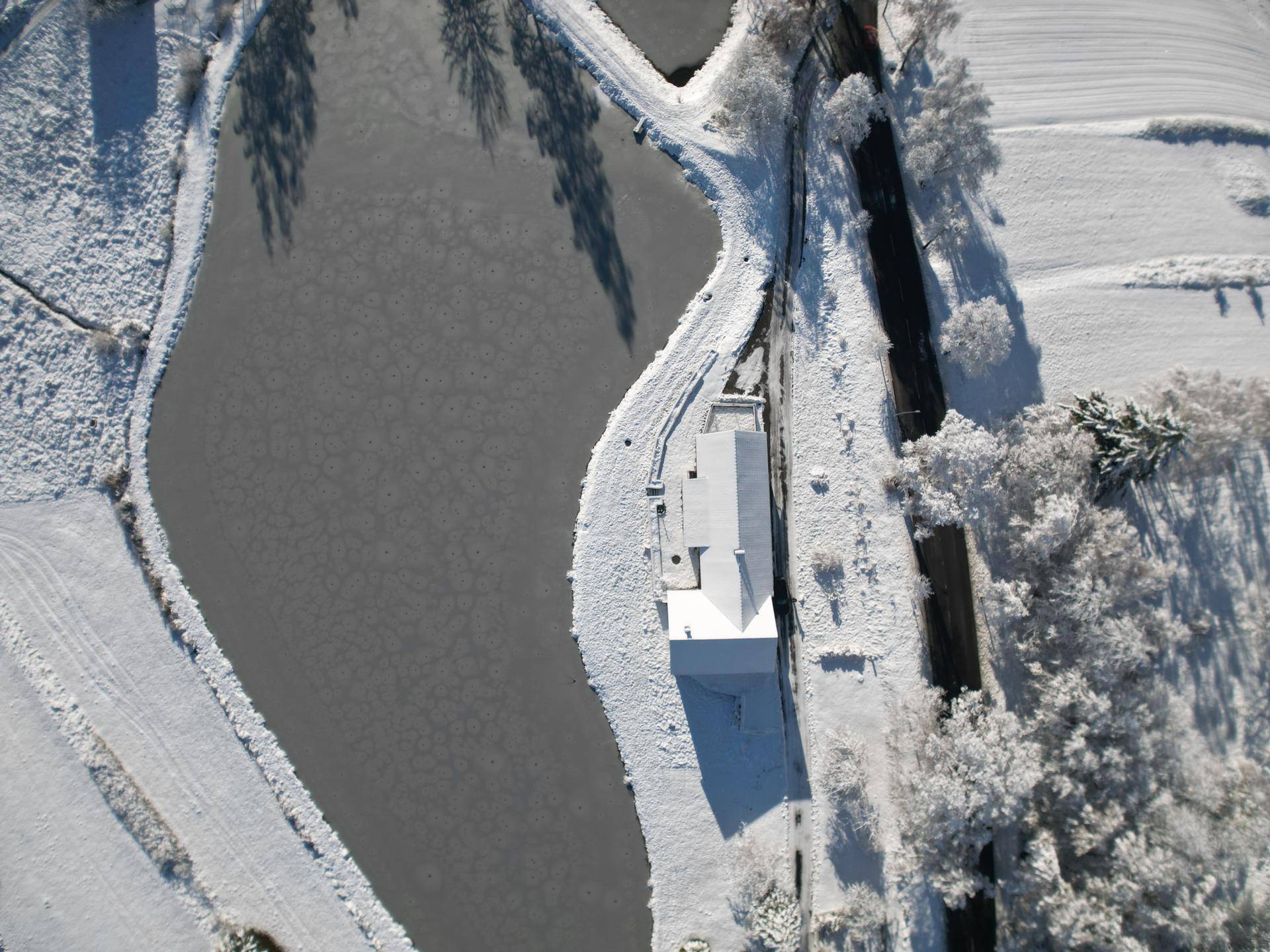 Luftaufnahme eines Hauses am zugefrorenen See, umgeben von verschneiter Winterlandschaft.