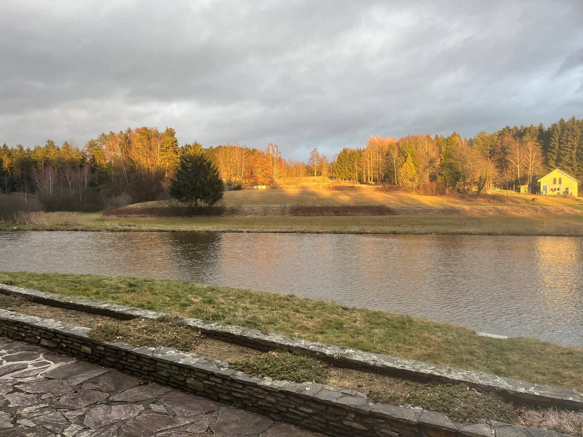 Malerischer Seeblick bei Sonnenuntergang oder Sonnenaufgang mit golden beleuchteten Bäumen und einem Haus in der Ferne.