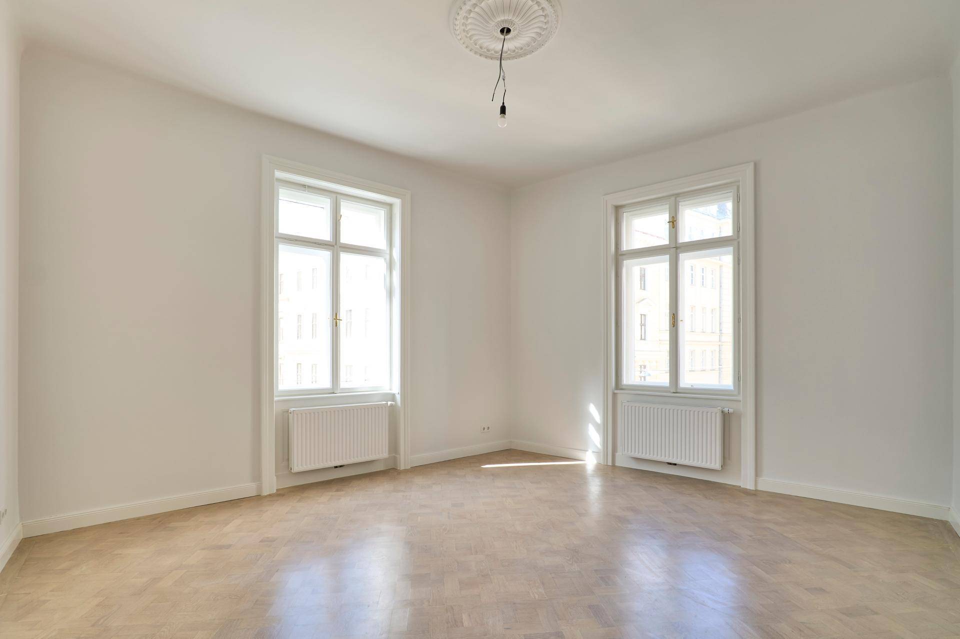 Heller Raum mit zwei hohen Fenstern, Fischgrätparkett und einer dekorativen Stuckrosette an der Decke.