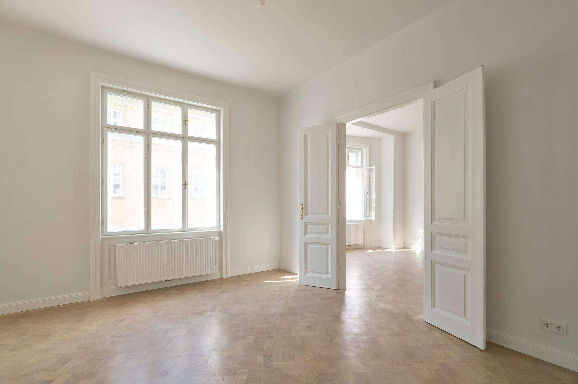 Geräumiges Zimmer mit großen Fenstern, hellem Fischgrätparkett und klassischen Flügeltüren zu angrenzenden Räumen.