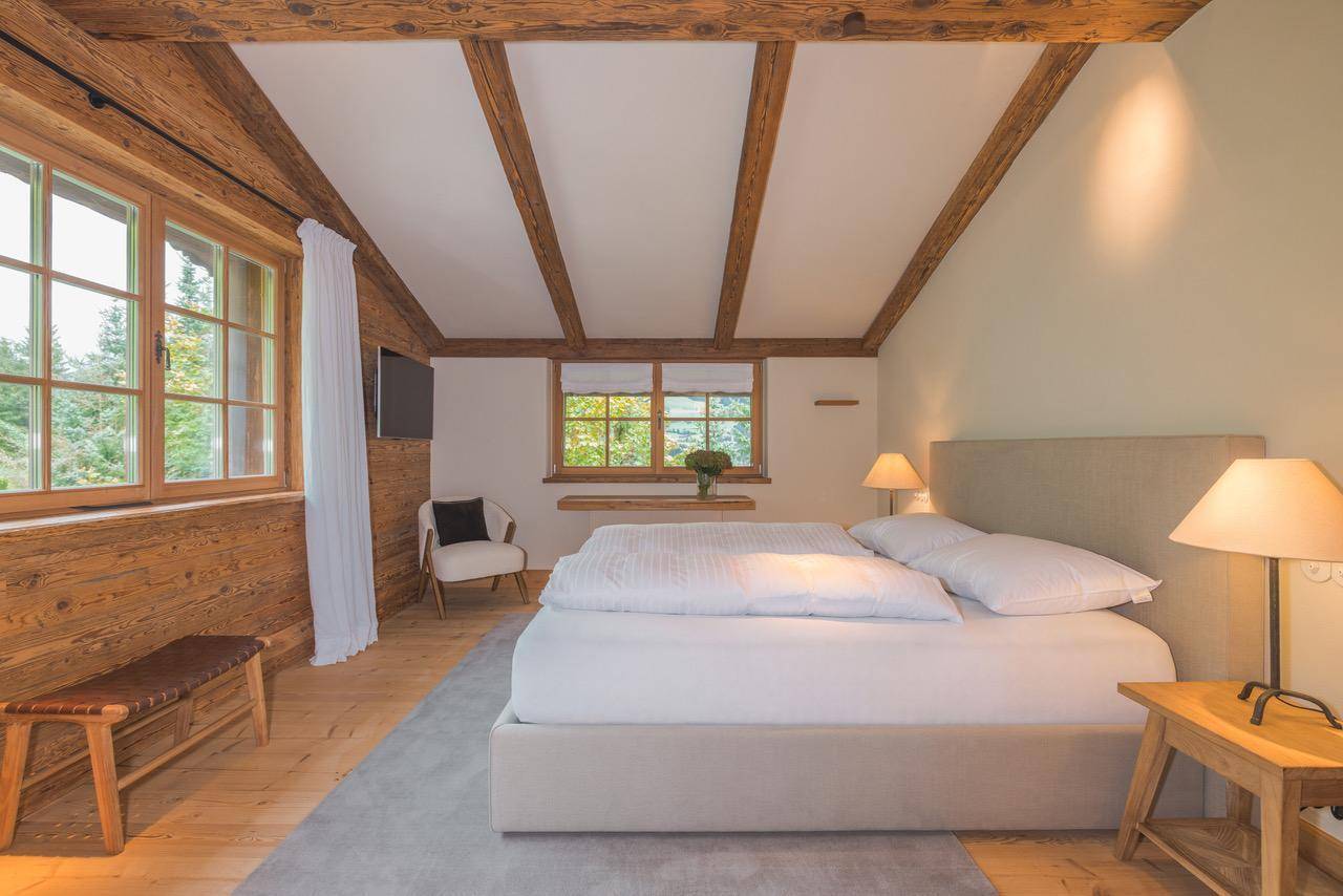 Rustikales Schlafzimmer mit Holzwänden, sichtbaren Deckenbalken und großen Fenstern mit Waldblick.