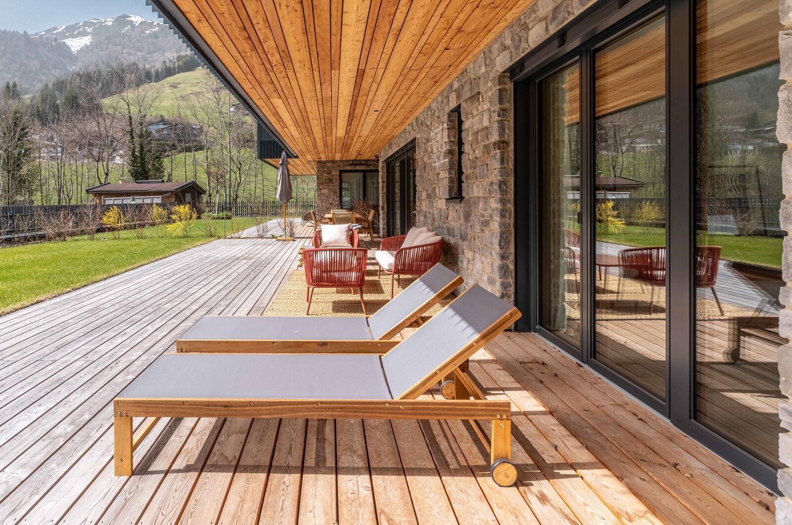 Große Holzterrasse mit Sonnenliegen und Sitzgelegenheiten, umgeben von grüner Landschaft.