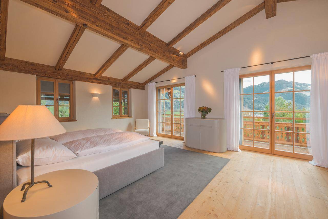 Lichtdurchflutetes Schlafzimmer mit Holzbalkendecke und weitem Blick auf die Berglandschaft vom Balkon.
