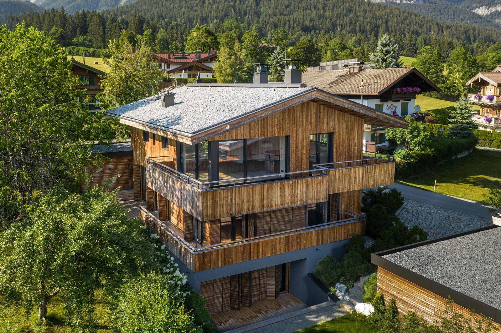 Luftaufnahme eines modernen Holzhauses mit mehreren Balkonen und Satteldach in grüner Landschaft.