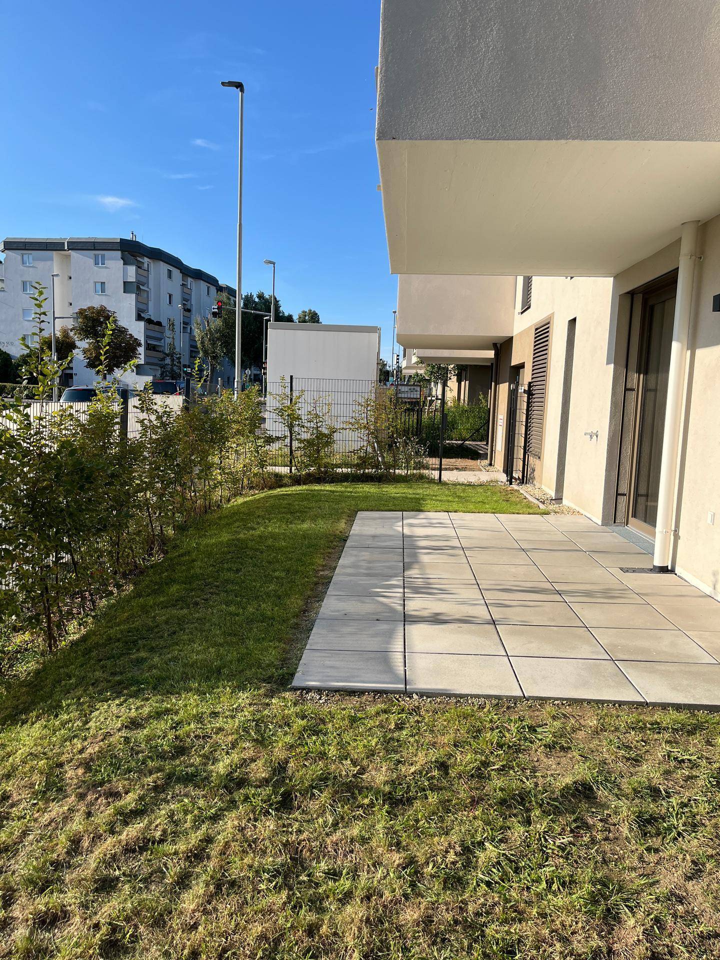 Sonniger Außenbereich mit gepflasterter Terrasse und angrenzendem Rasen, umgeben von Grün.