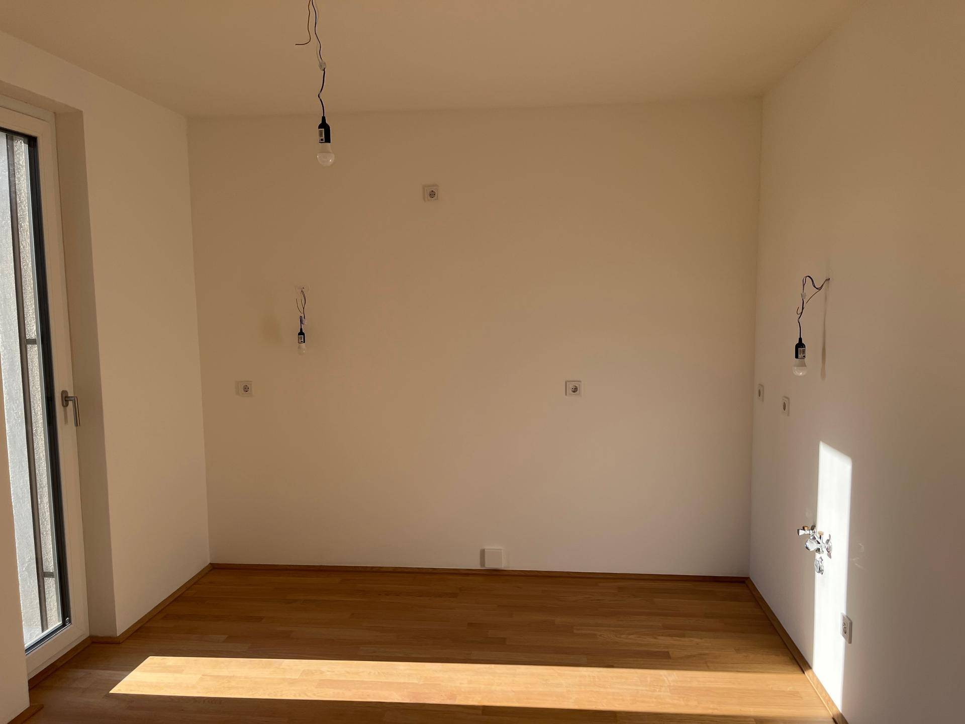 Leerer Küchenbereich mit Parkettboden und Anschlüssen, bereit für die Installation einer Einbauküche.