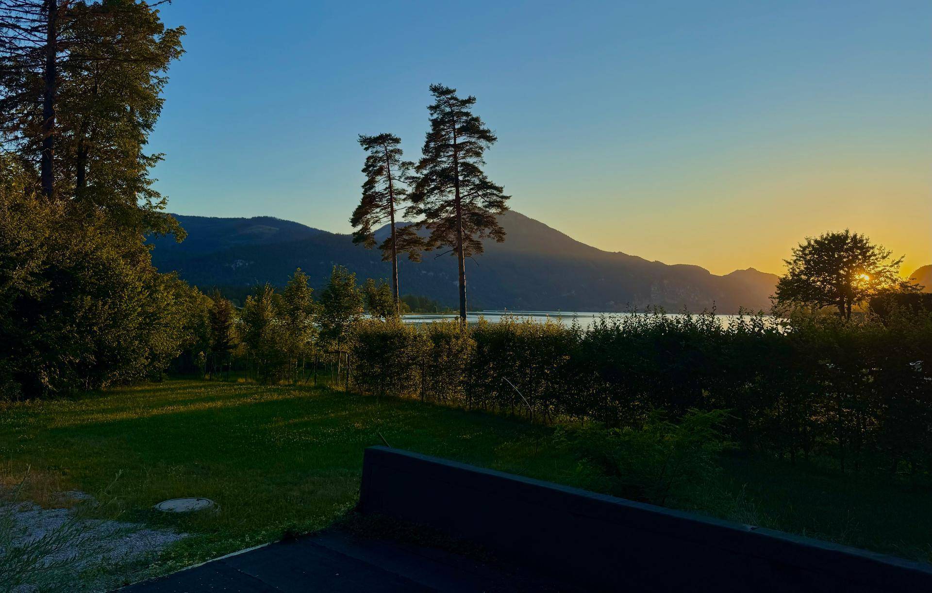 Idyllischer Sonnenuntergang über einem See mit majestätischen Bergen im Hintergrund, umgeben von üppigem Grün und einem gepflegten Garten.