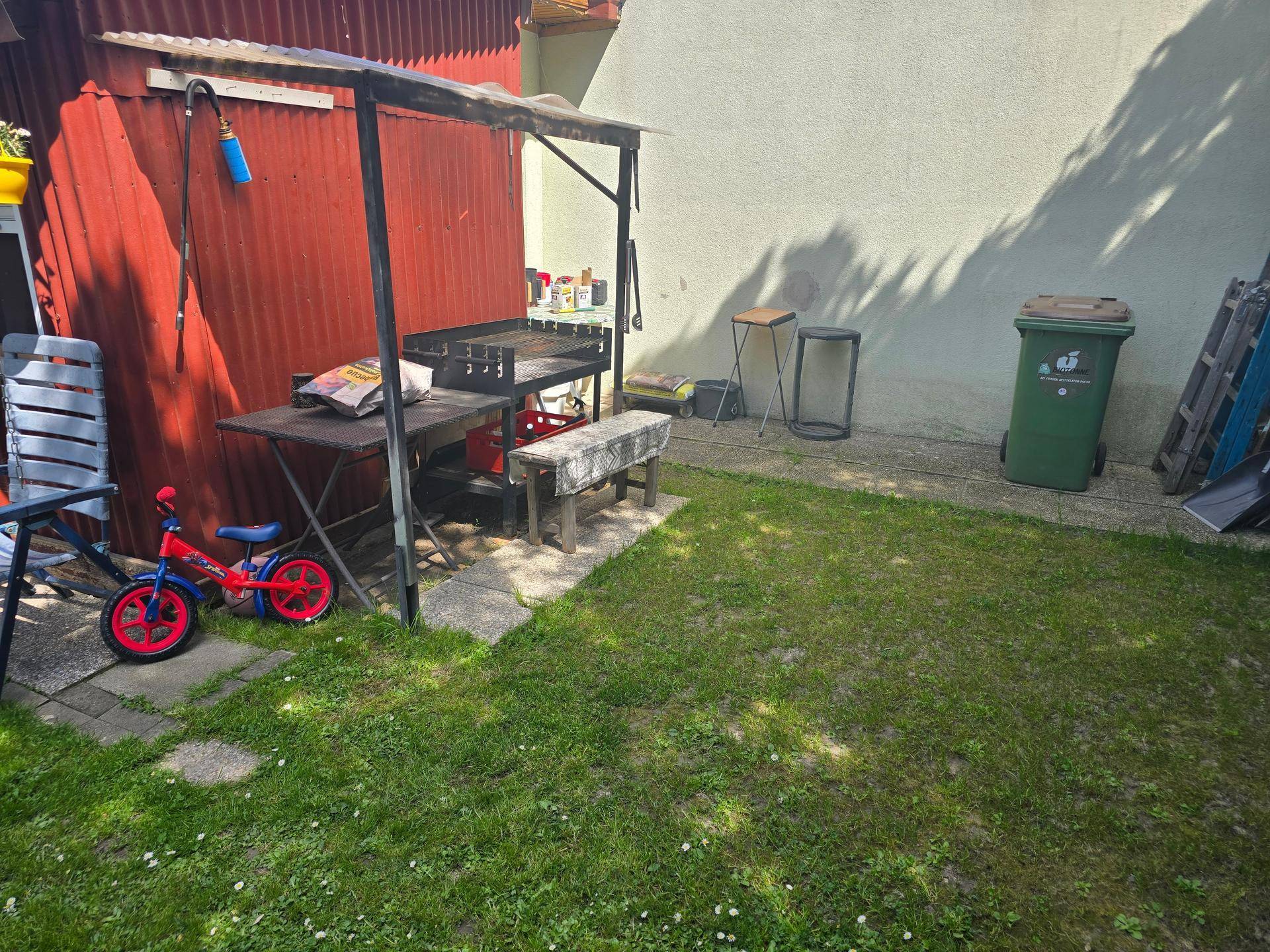 Gartenbereich mit Grillplatz, Rasenfläche und einem kleinen Kinderfahrrad.