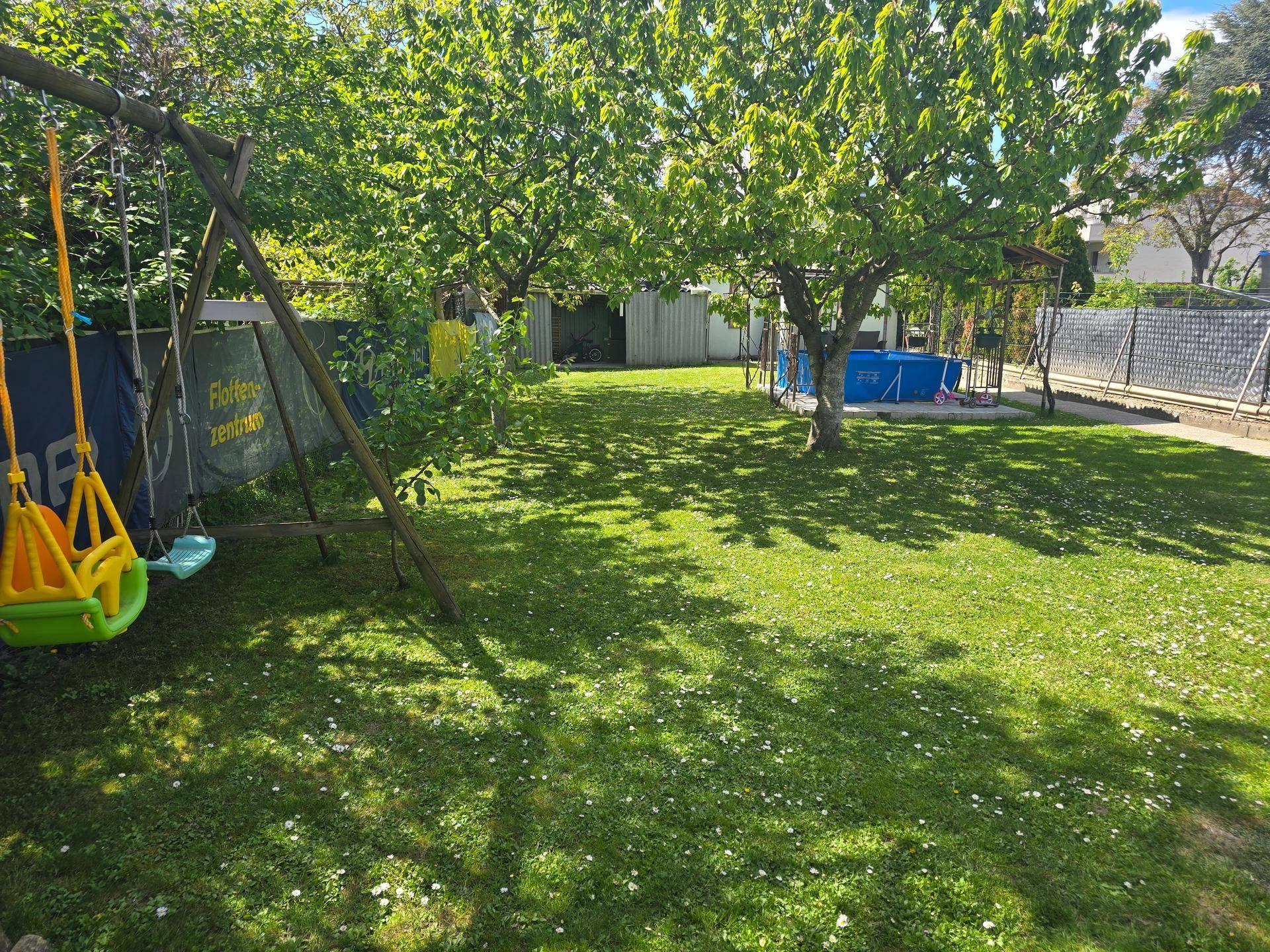 Weitläufiger Garten mit Schaukel, Bäumen und einem Aufstellpool im Hintergrund.