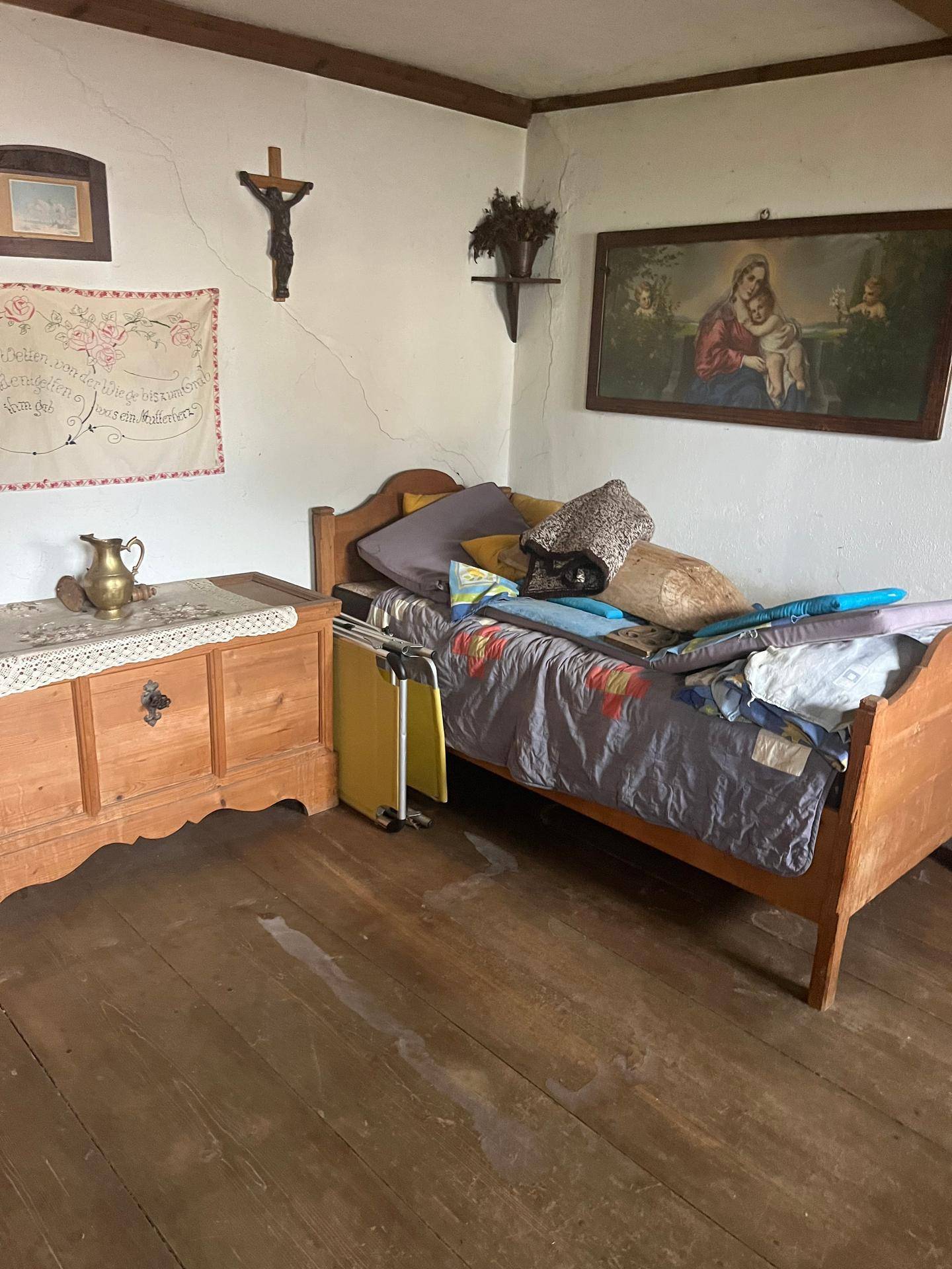 Einfaches Schlafzimmer mit Holzbett, Holzboden und religiösen Dekorationen an der Wand, hell und freundlich.