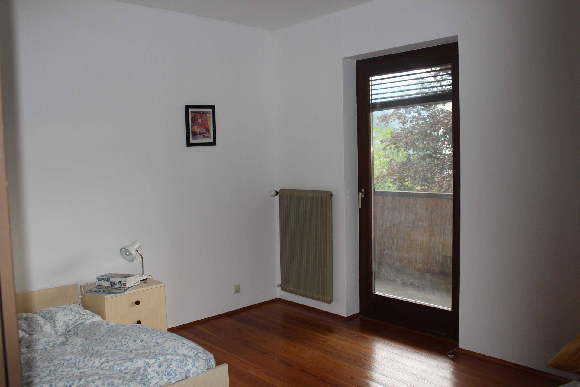 Schlichtes Schlafzimmer mit Holzboden, Heizkörper und direktem Zugang zum Balkon.