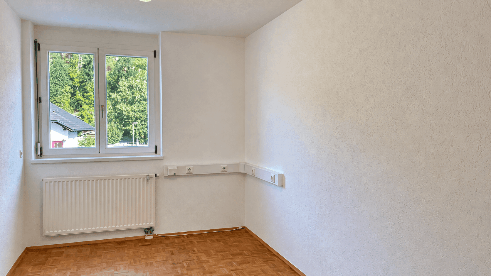 Freundliches Zimmer mit Holzboden, großen Fenstern und einer angenehmen Aussicht ins Grüne.