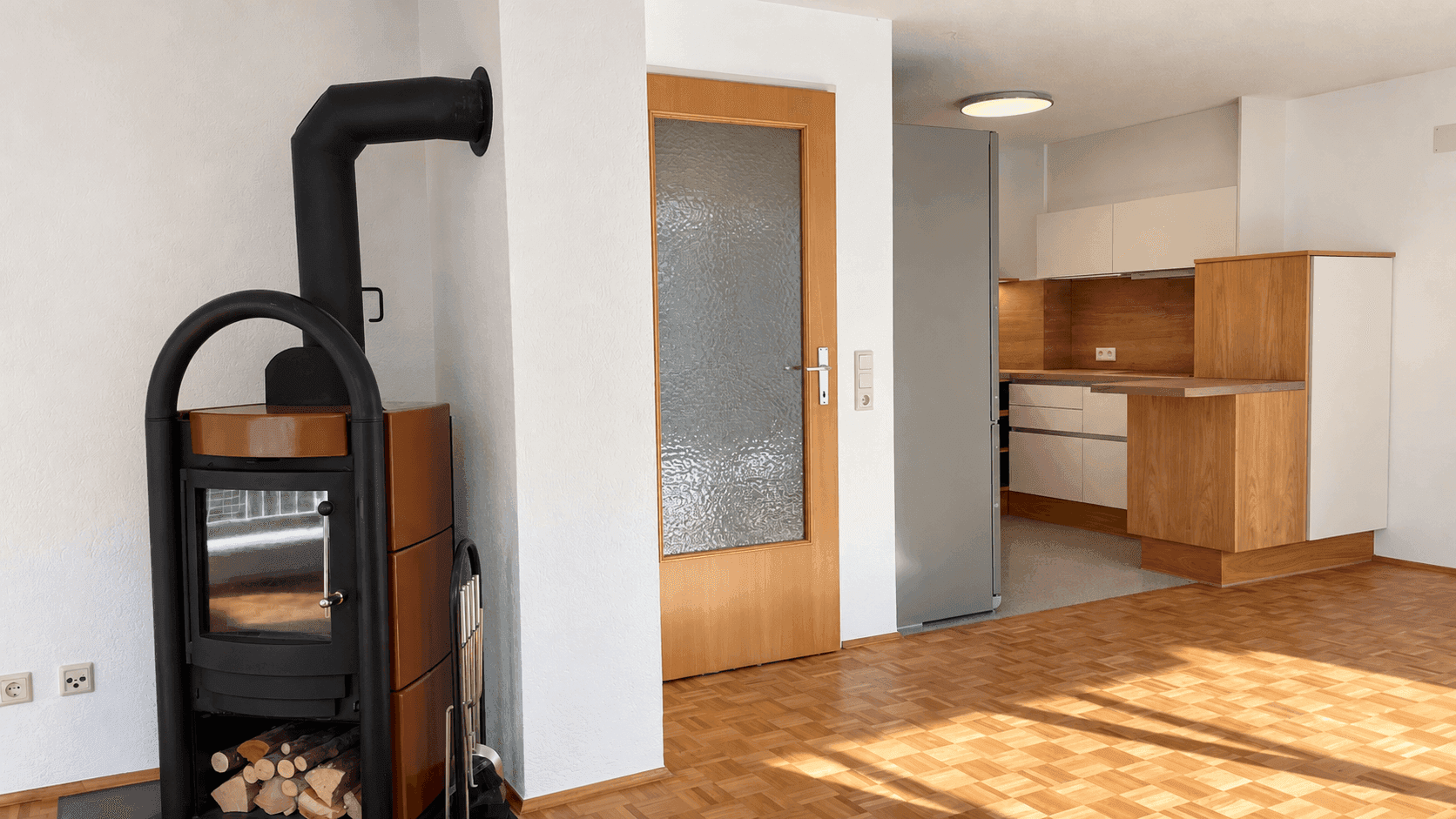 Offener Wohnbereich mit Holzofen und Blick auf die moderne, helle Küche mit Holzelementen.
