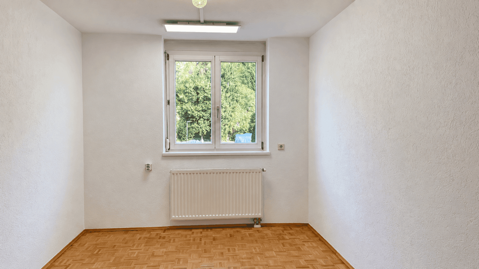 Helles Zimmer mit Parkettboden, weiß gestrichenen Wänden und einem Fenster mit Blick ins Grüne.