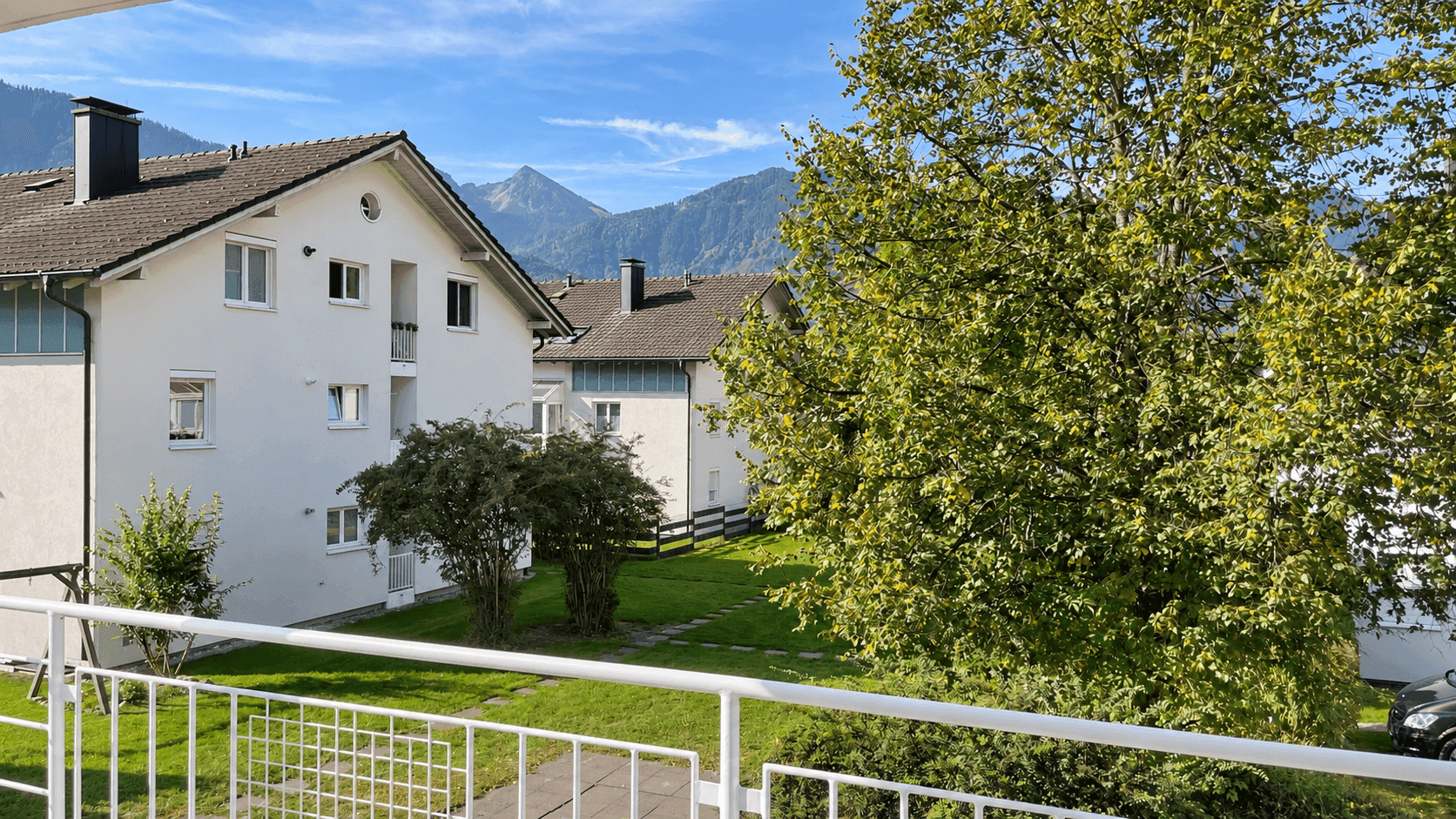 Panoramablick von einem Balkon auf Wohngebäude, gepflegte Grünflächen und majestätische Berge.