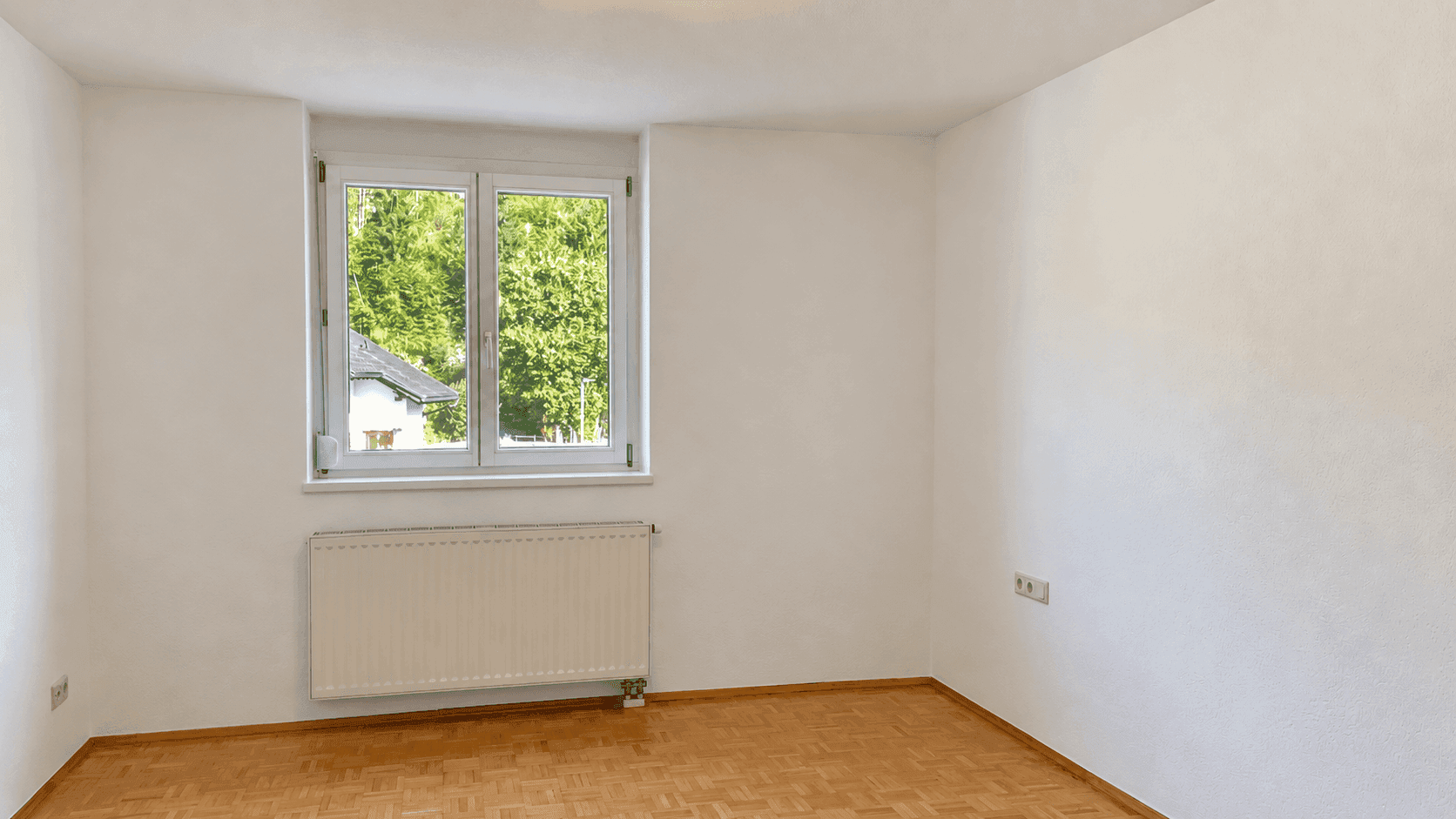 Leeres Zimmer mit Parkettboden, weißer Wandfarbe und einem Fenster, das viel Tageslicht hereinlässt.