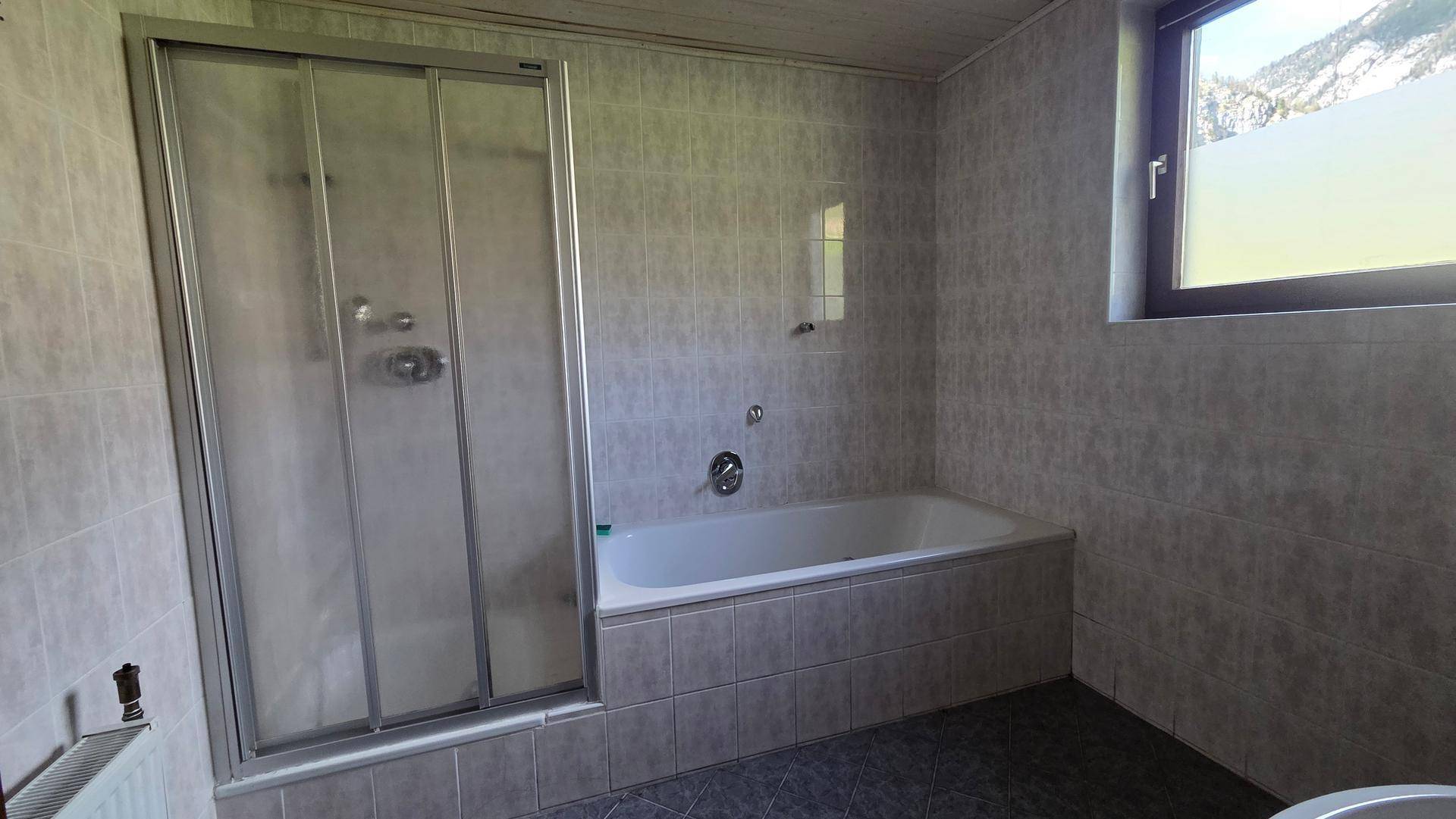 Gefliestes Badezimmer mit Badewanne, Duschkabine und Fenster, im Stil der 80er Jahre.