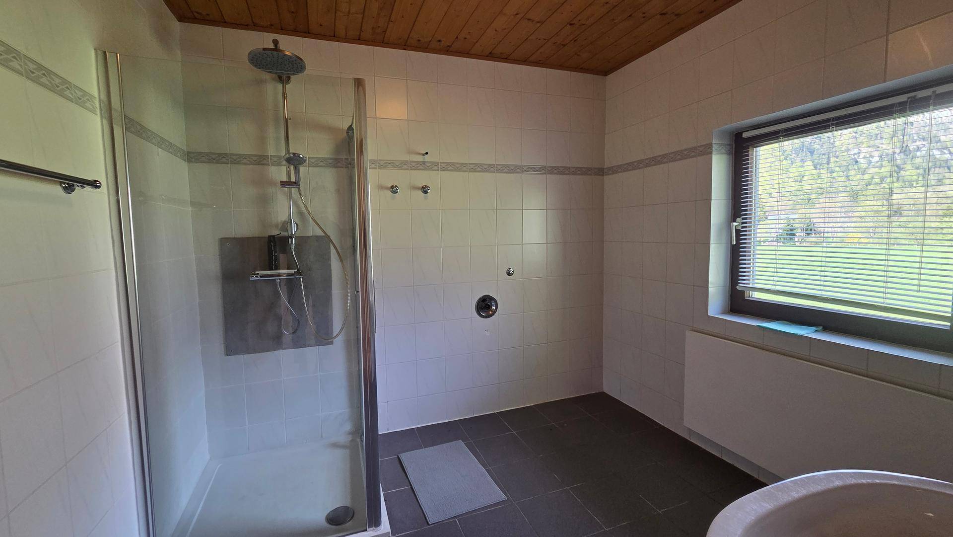 Badezimmer mit Duschkabine, Fliesenboden und Fenster mit Blick ins Grüne.
