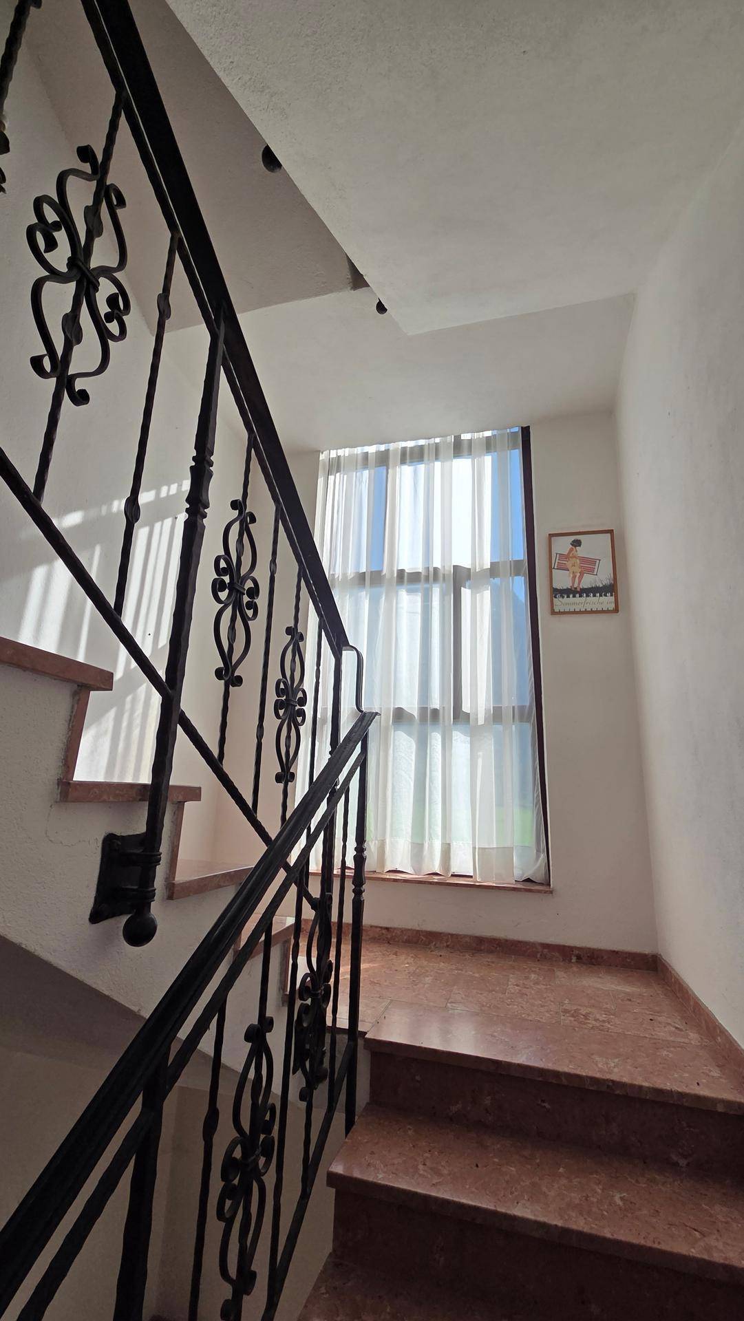 Elegantes Treppenhaus mit schmiedeeisernem Geländer, Marmorstufen und einem großen Fenster mit Vorhängen.