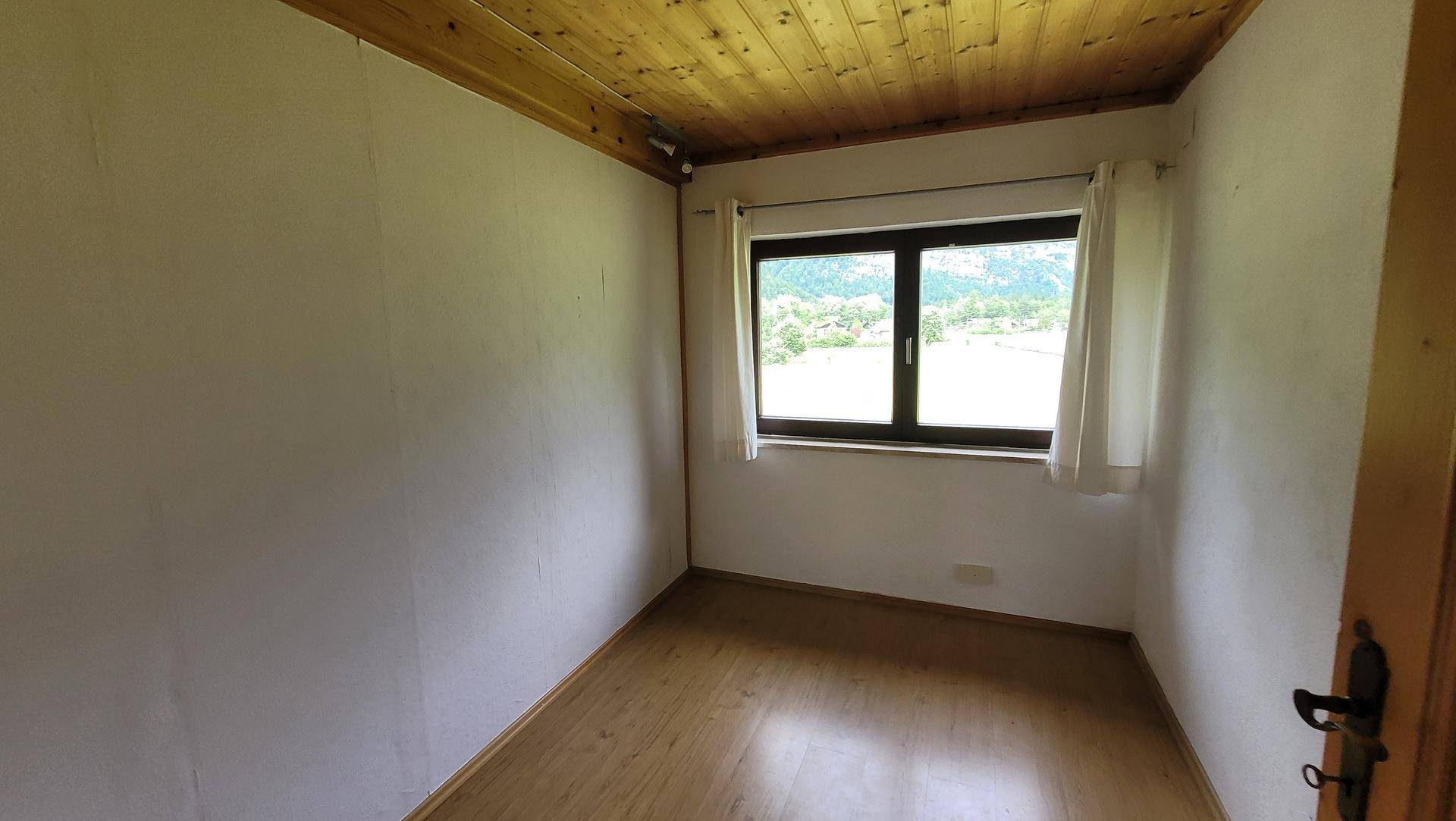 Helles, leeres Zimmer mit Holzboden und Fenster mit Blick auf die grüne Umgebung.