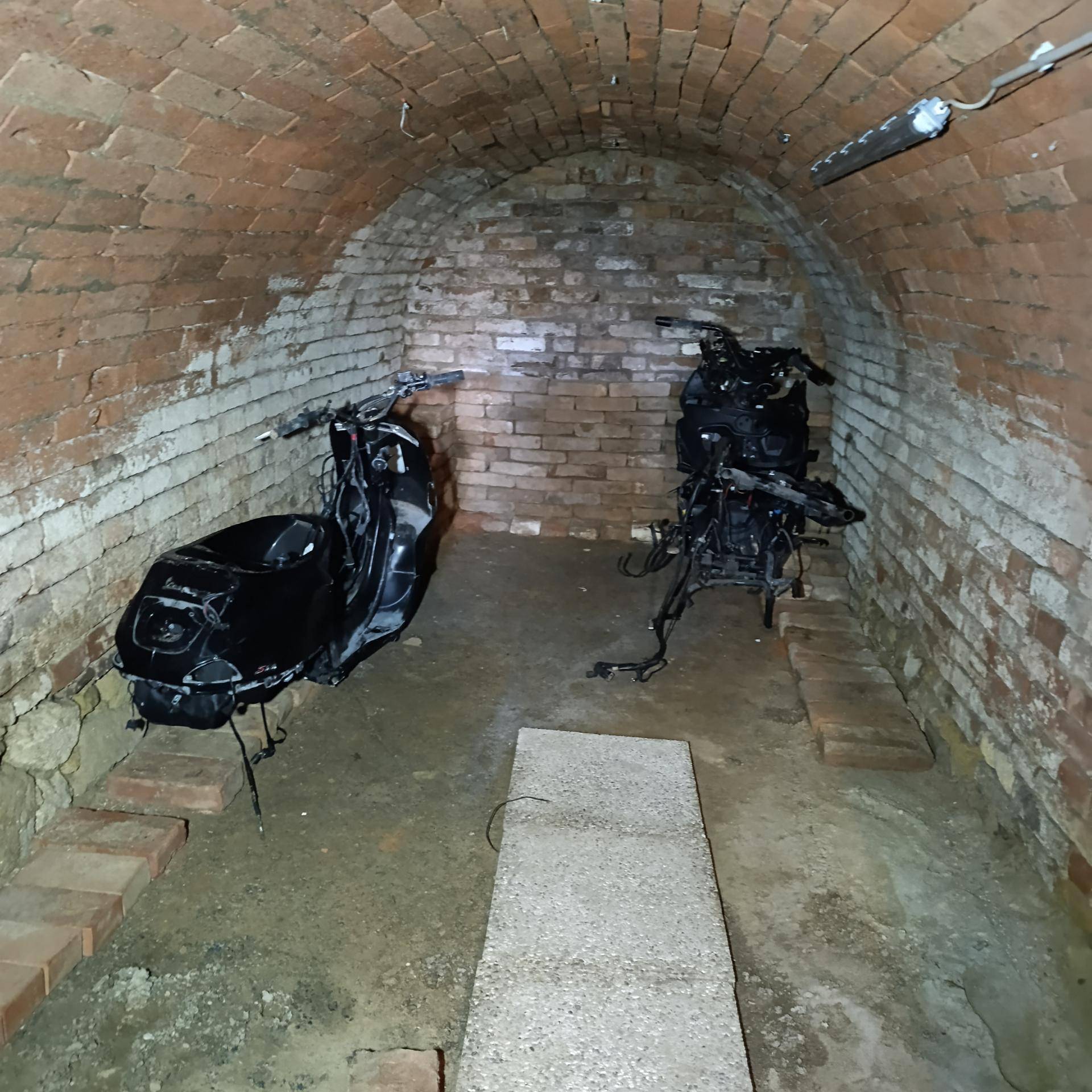 Ein gewölbter Ziegelkeller, der als Lagerraum für zwei Motorräder dient, mit einem Betonweg in der Mitte.