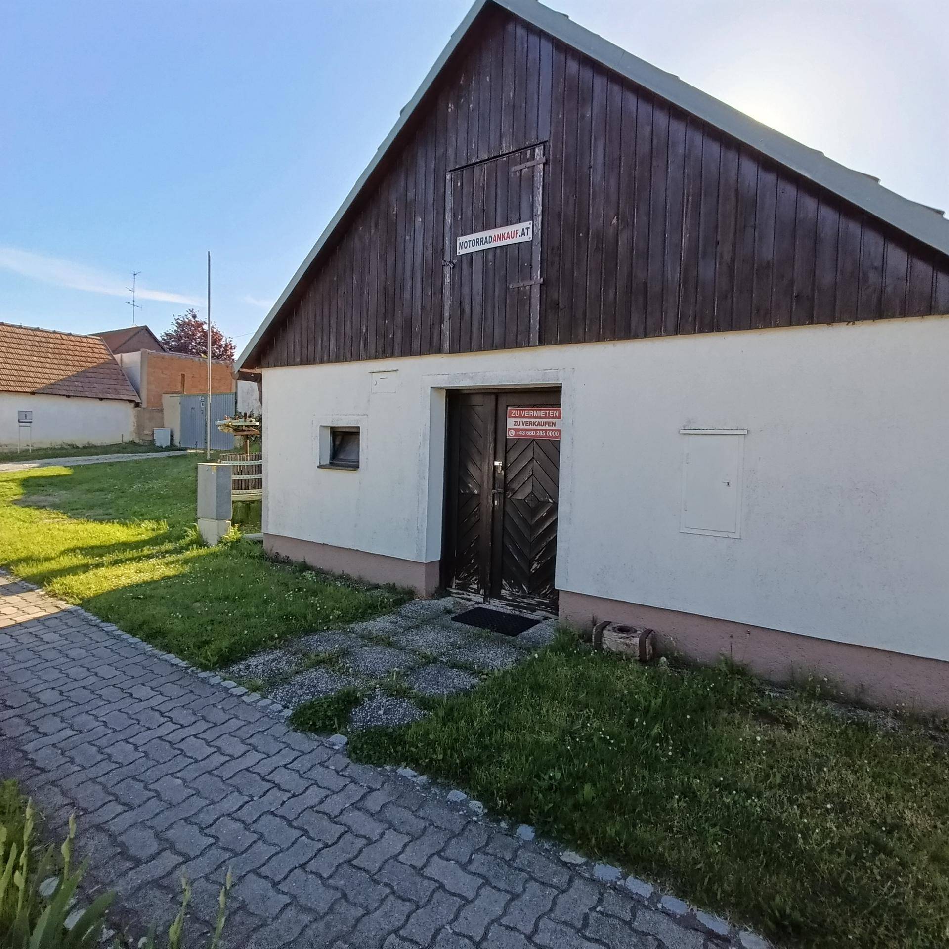 Nahaufnahme der Gebäudefassade mit Holztüren und einem kleinen Fenster, umgeben von Grünflächen und einem gepflasterten Weg.
