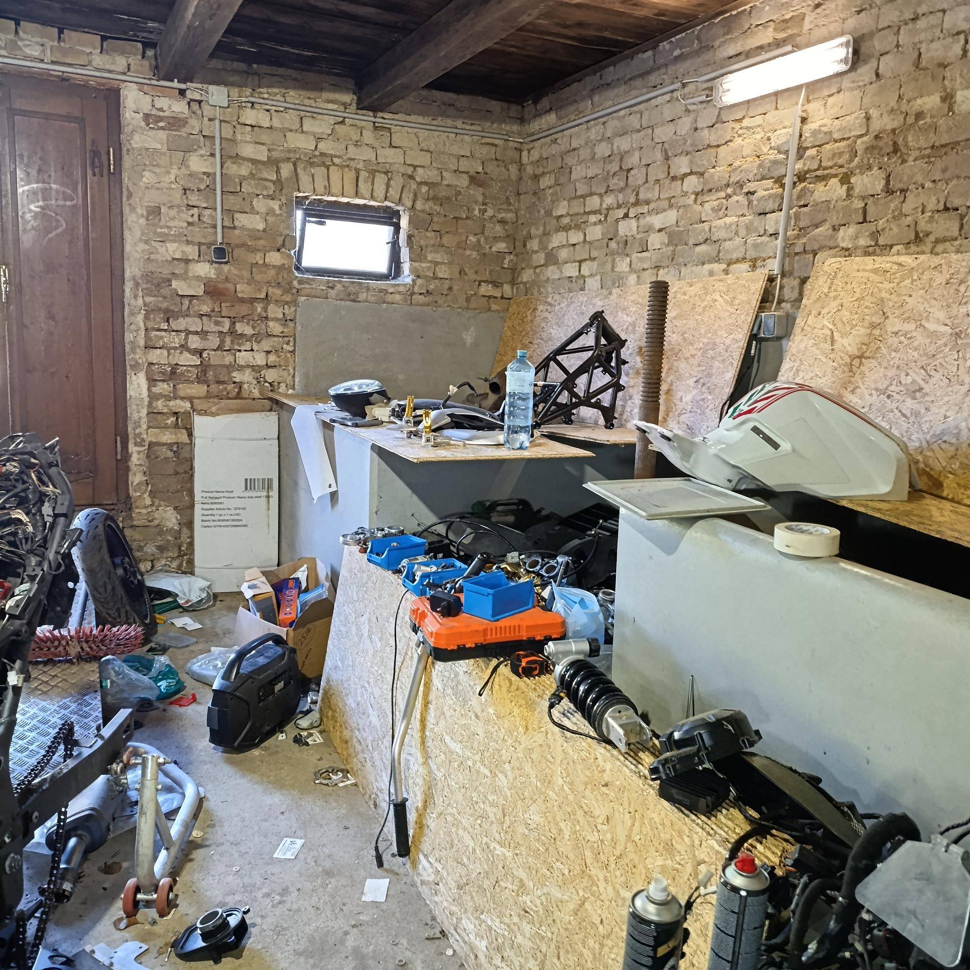 Ein unaufgeräumter Werkstattraum mit Werkbänken aus Holzplatten und verschiedenen Motorradteilen und Werkzeugen.