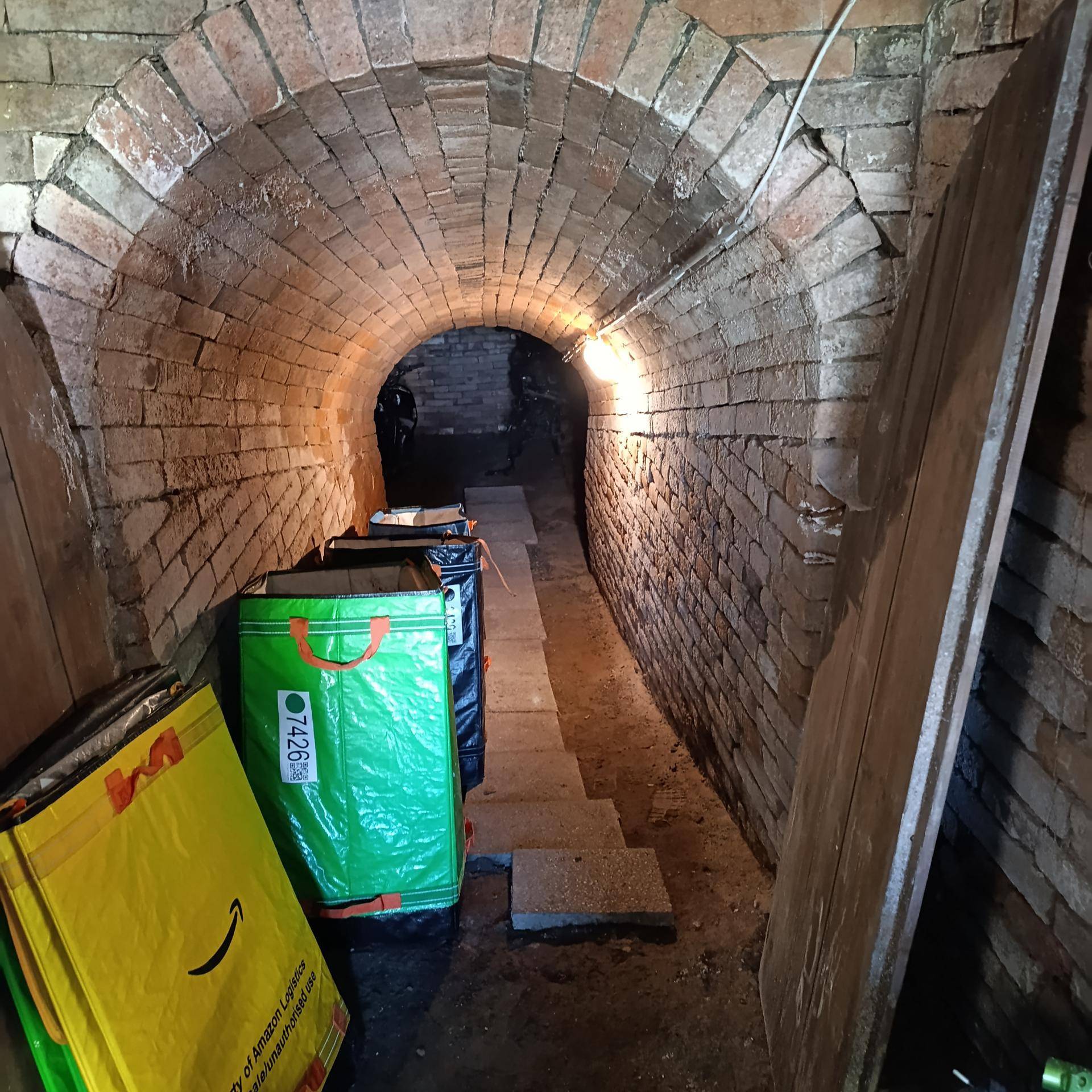 Ein langer, gewölbter Ziegelkeller mit einer einzelnen Glühbirne und einigen Lagerbehältern entlang der Wand.
