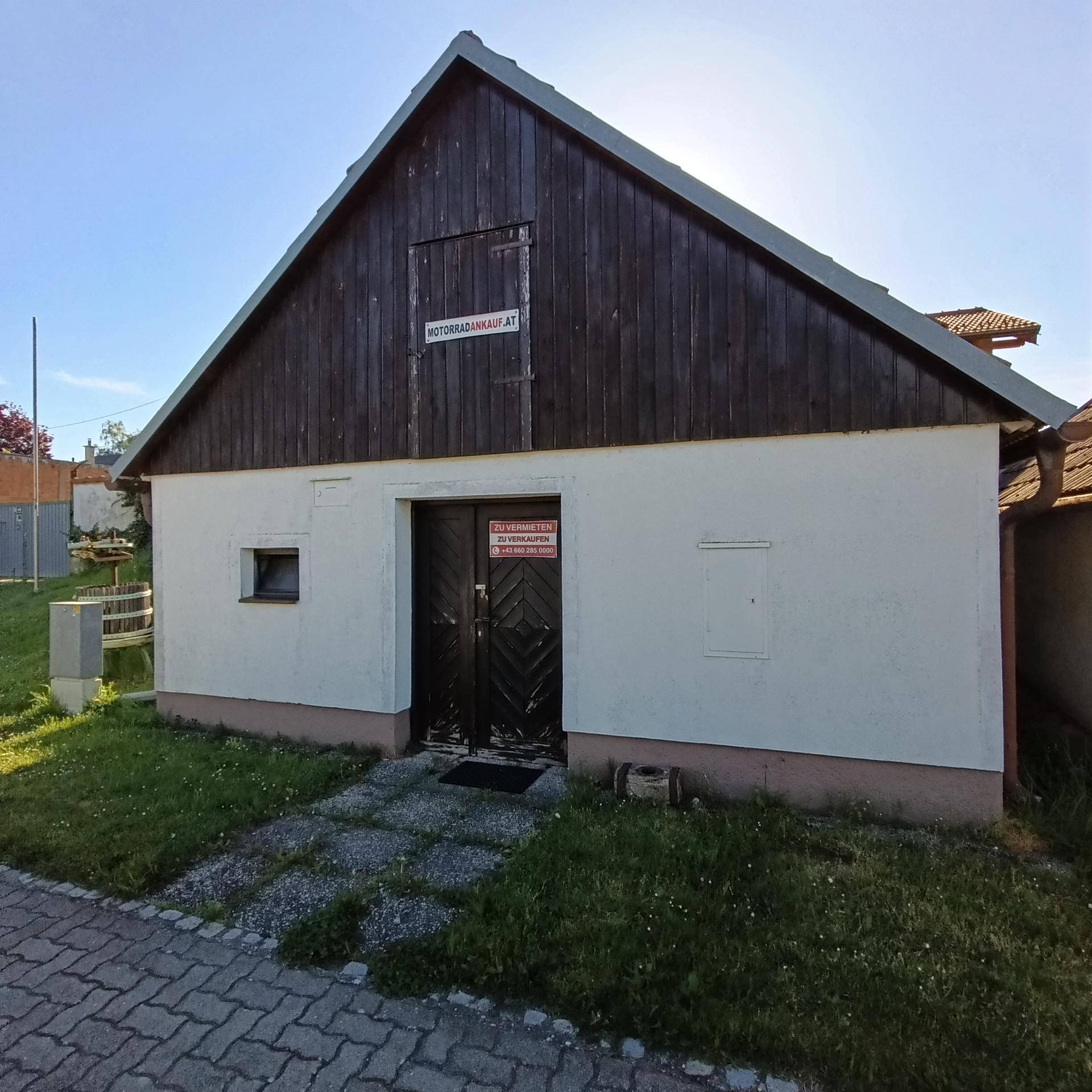 Seitenansicht eines kleinen Gebäudes mit Holztüren und einem Schild an der Fassade, umgeben von Grasflächen.