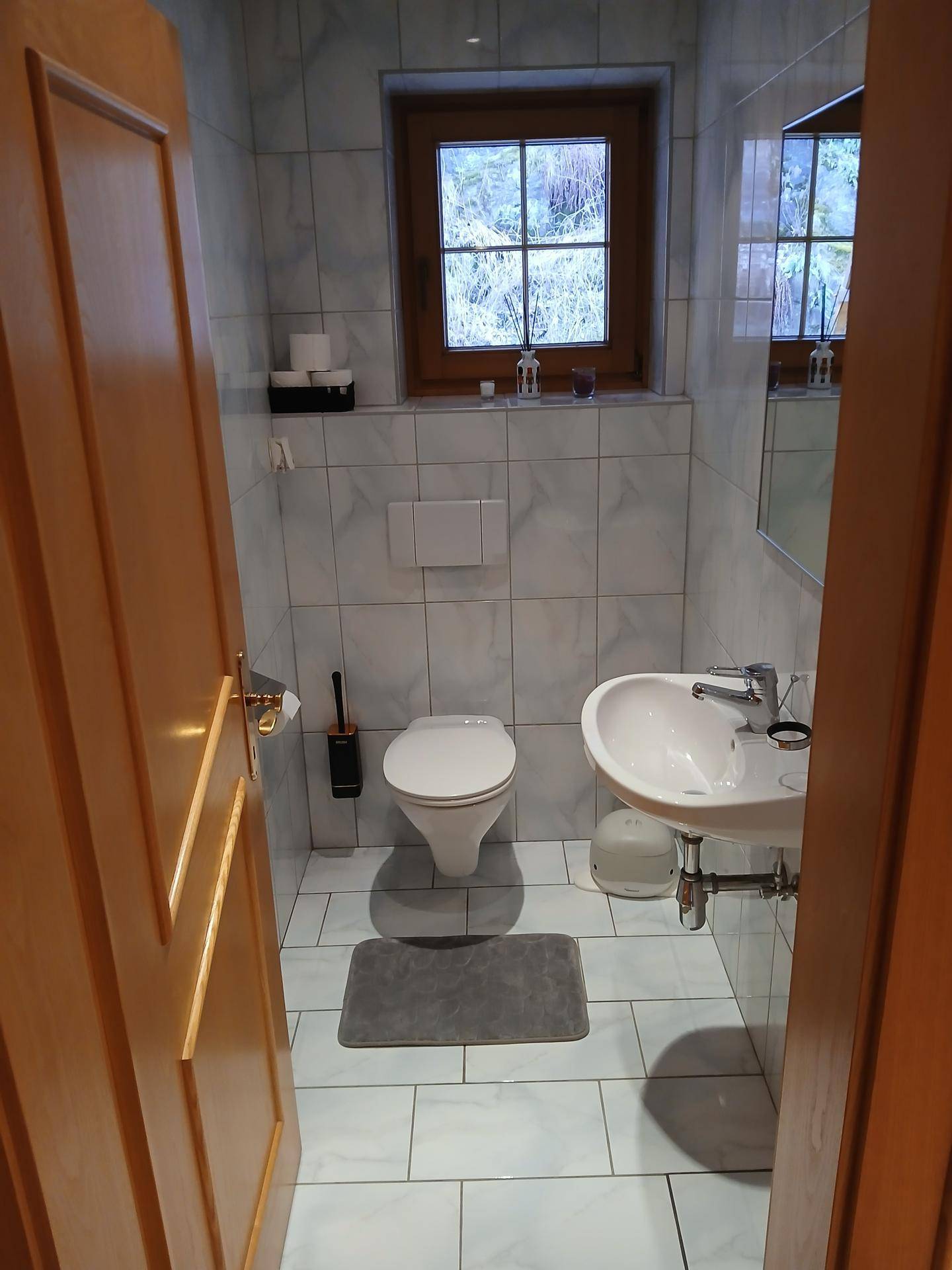 Kleines Gäste-WC mit Fenster, Wand-WC und Waschbecken in hellen Fliesen.