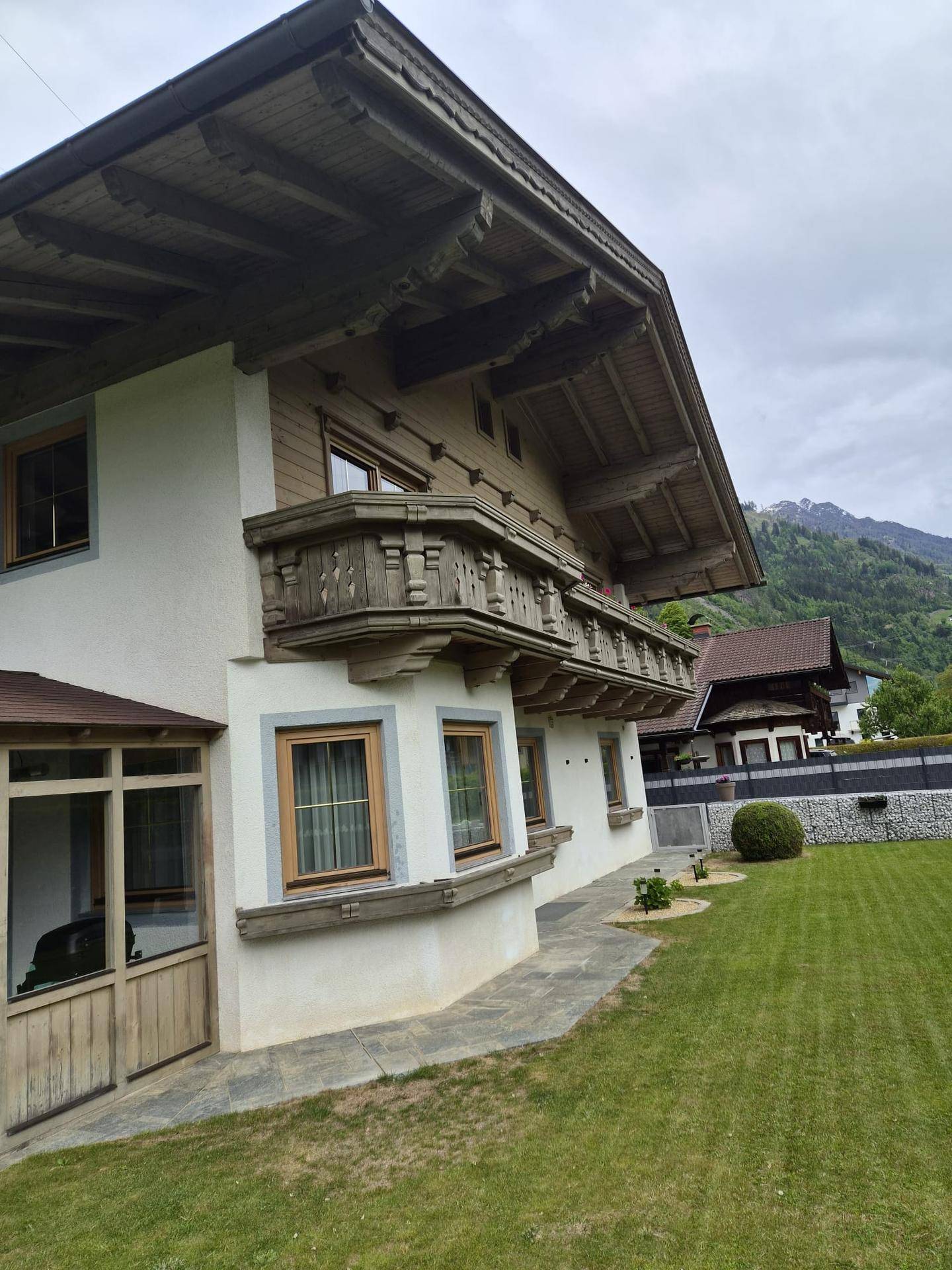 Seitenansicht eines traditionellen Alpenhauses mit großem Rasenbereich und Bergblick unter bewölktem Himmel.