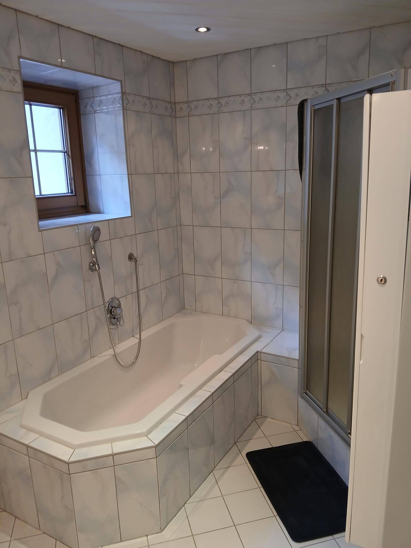Badezimmer mit Eckbadewanne, separater Duschkabine und Fenster in hellen Fliesen.