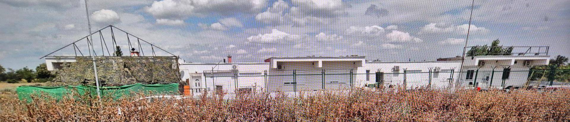 Panoramablick auf ein weißes Gebäudeensemble mit Zaun und trockenen Feldern im Vordergrund.